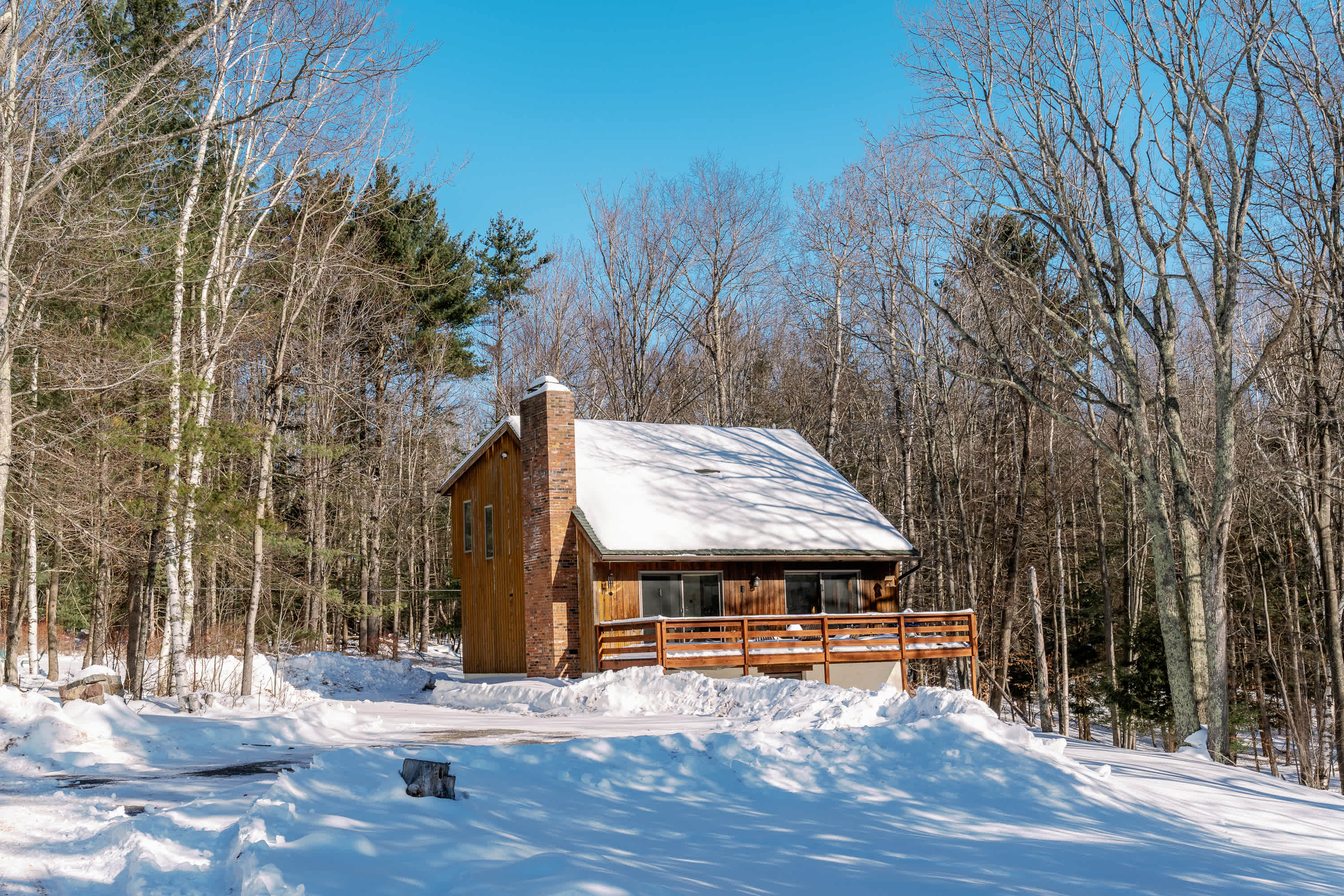 A rustic wooden cabin with a brick chimney and a snow-covered roof, surrounded by bare trees and a snowy driveway under a clear blue sky. A wooden deck extends from the front of the house.