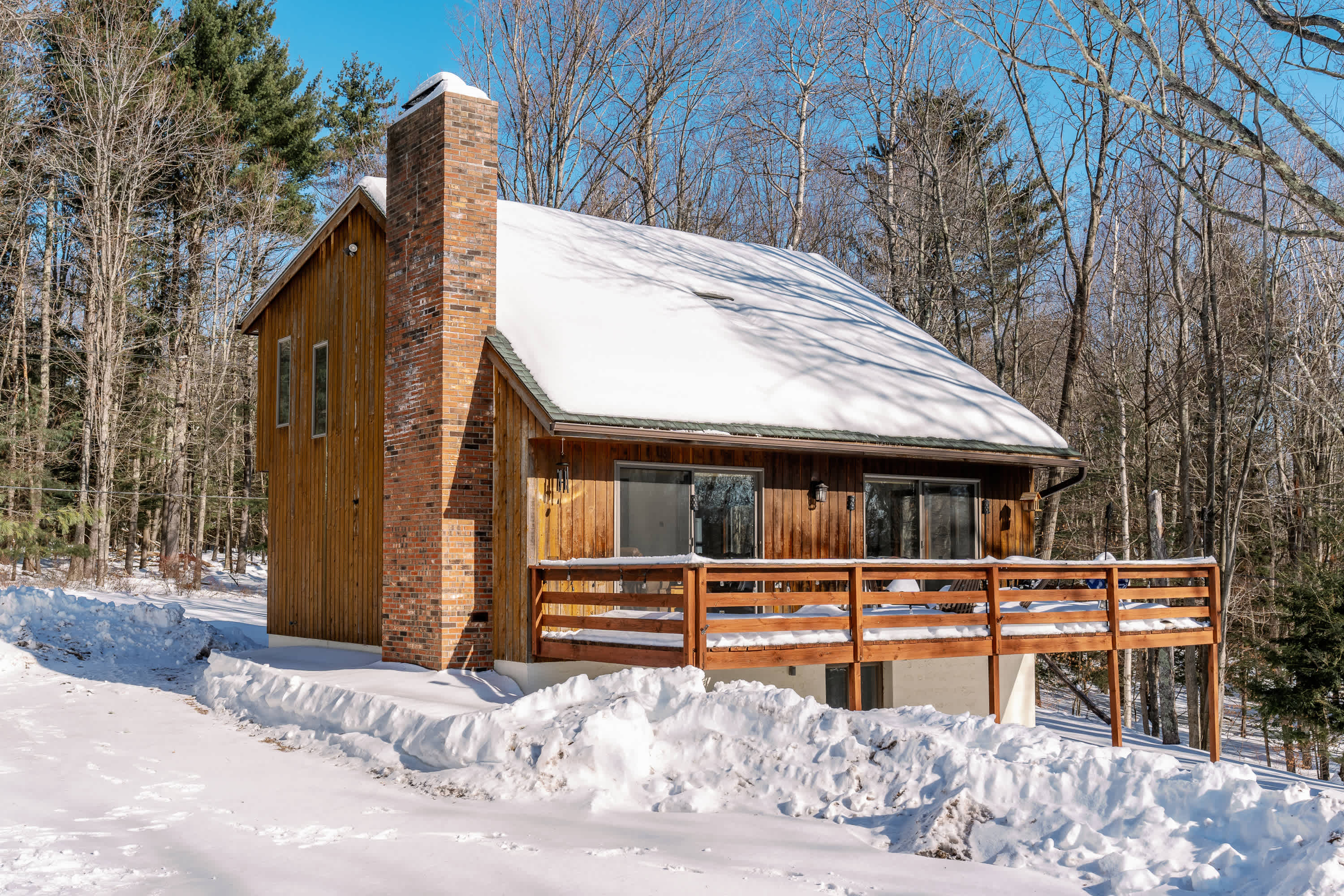 A charming wooden cabin with a snow-covered roof and a large brick chimney stands amidst a snowy landscape, surrounded by bare winter trees. A spacious wooden deck with railings extends from the front of the house.