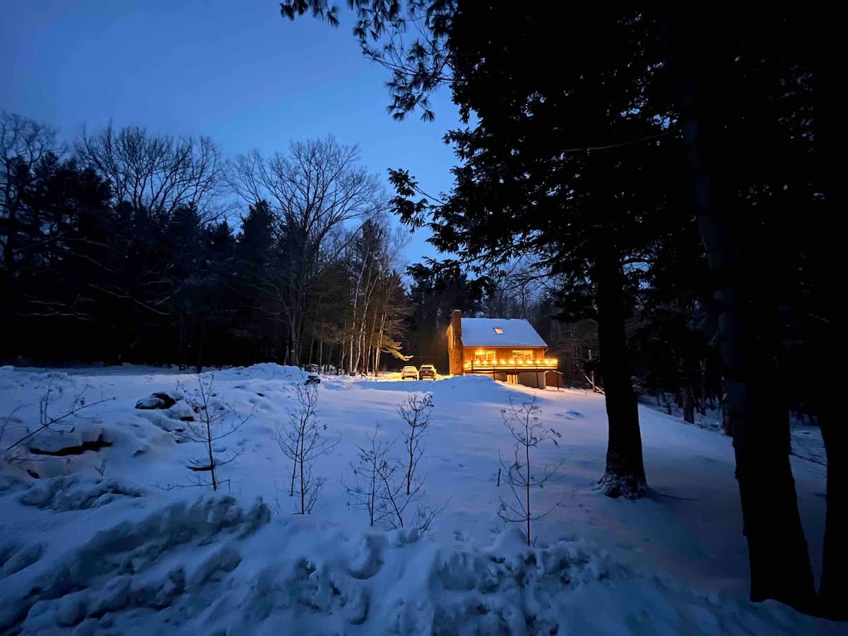 A charming cabin illuminated at dusk, surrounded by deep snow and bare trees under a dark blue sky. The warm glow from the windows contrasts with the cold, snowy landscape.