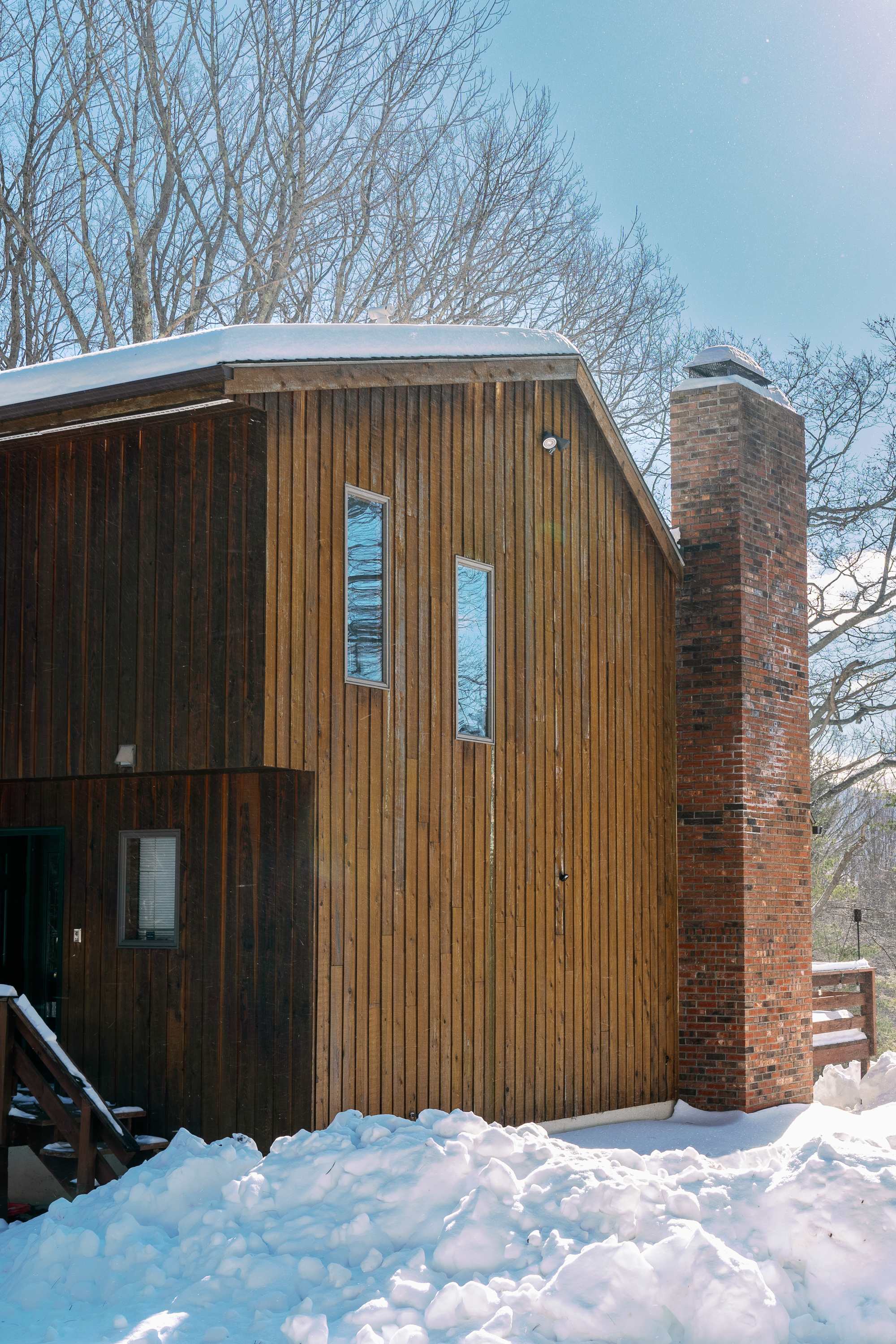 The side of a rustic wooden cabin with vertical wood siding and a prominent brick chimney, surrounded by deep snowdrifts and bare winter trees under a bright sky. Several windows are visible on the wooden facade.