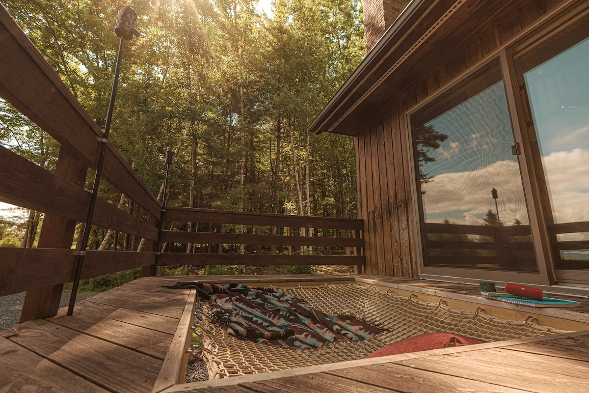 A wooden deck features a built-in net hammock filled with colorful blankets and pillows, overlooking a lush green forest under bright sunlight. A sliding glass door leads into the house, and a tiki torch stands nearby.