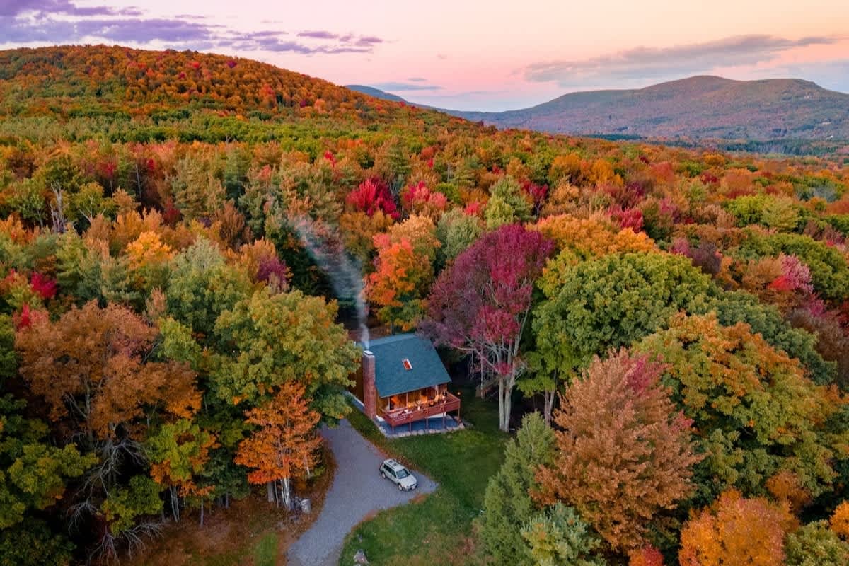 An aerial view showcases a charming cabin nestled amidst a vibrant forest of autumn trees, with a winding driveway and distant mountains under a colorful sunset sky.