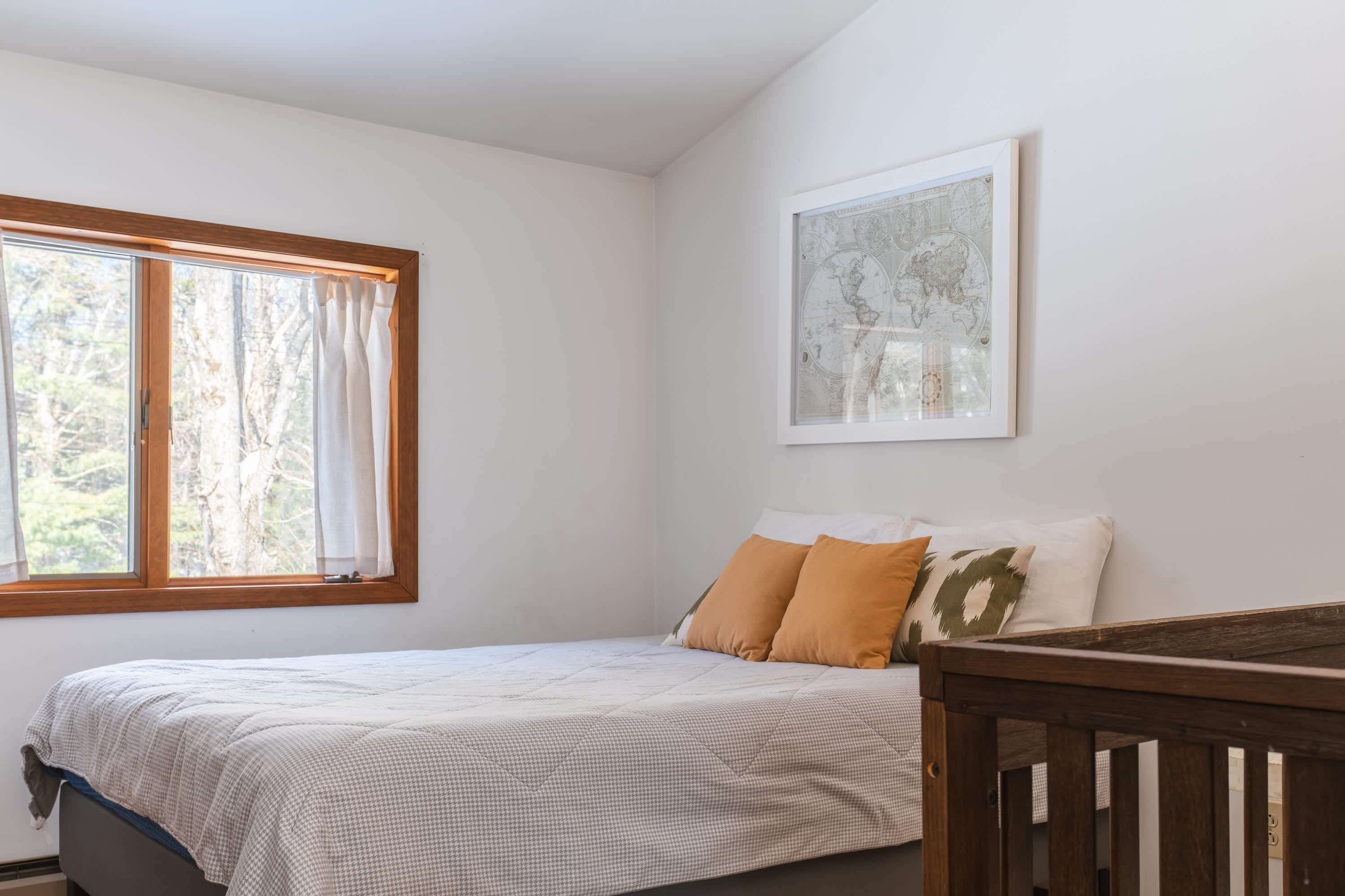 Another view of the bedroom with a queen bed and a dark wood crib, featuring a large window with white curtains. The room has hardwood floors and a world map art piece.