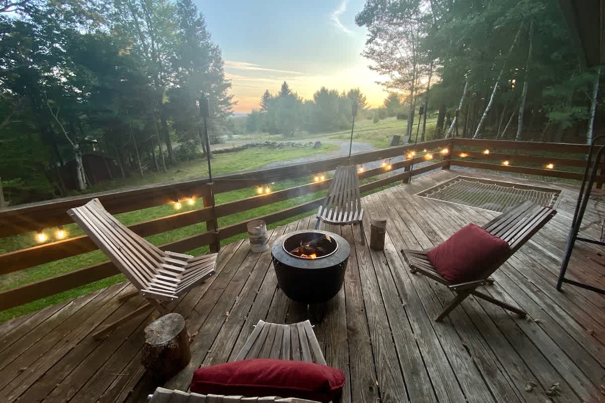 A spacious wooden deck furnished with several slatted wooden chairs, a central fire pit, and string lights overhead. The deck overlooks a grassy field and distant trees under a warm sunset sky.