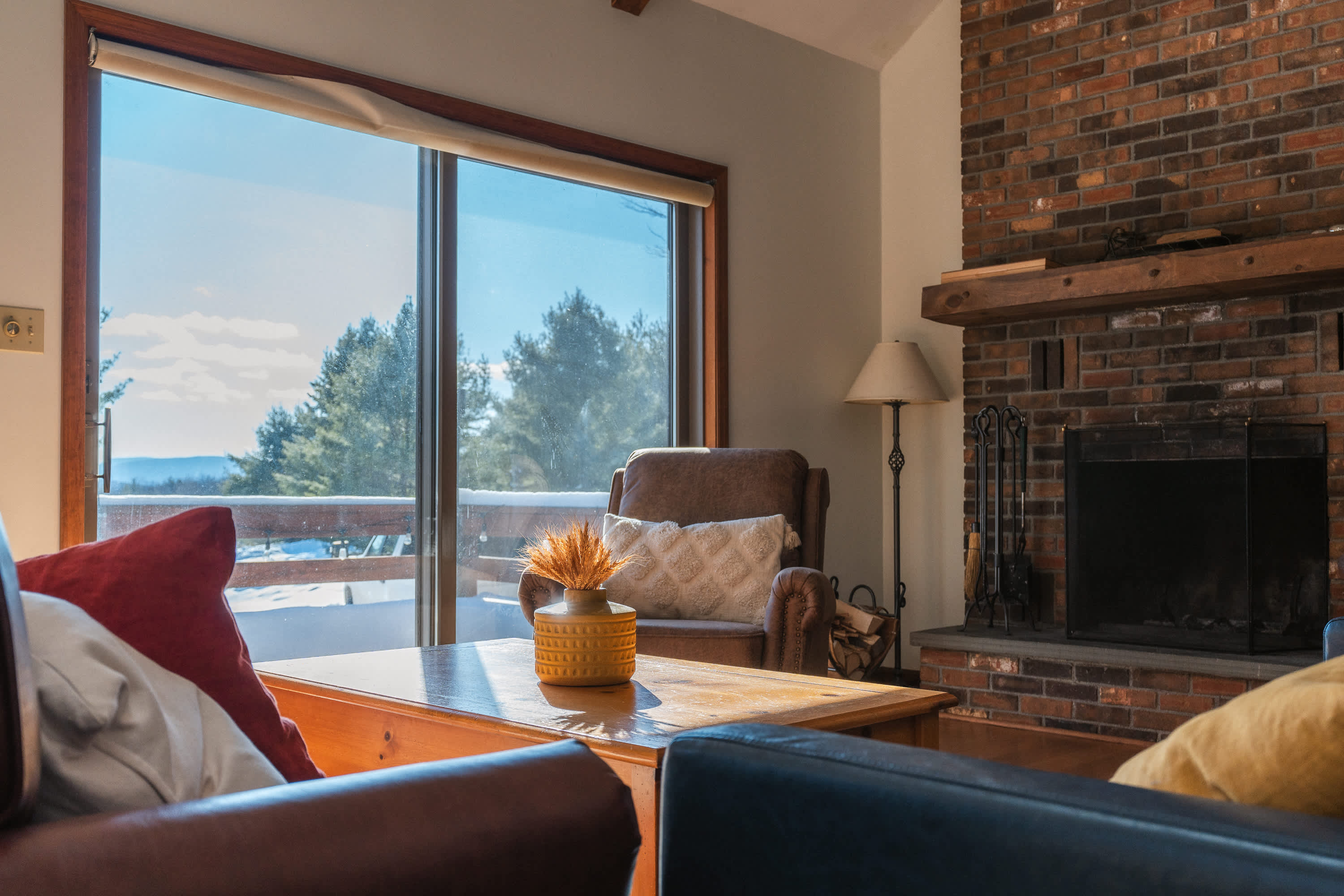 A cozy living room features a large brick fireplace with a wooden mantel, an armchair, and a coffee table, with a large window offering a snowy mountain view.