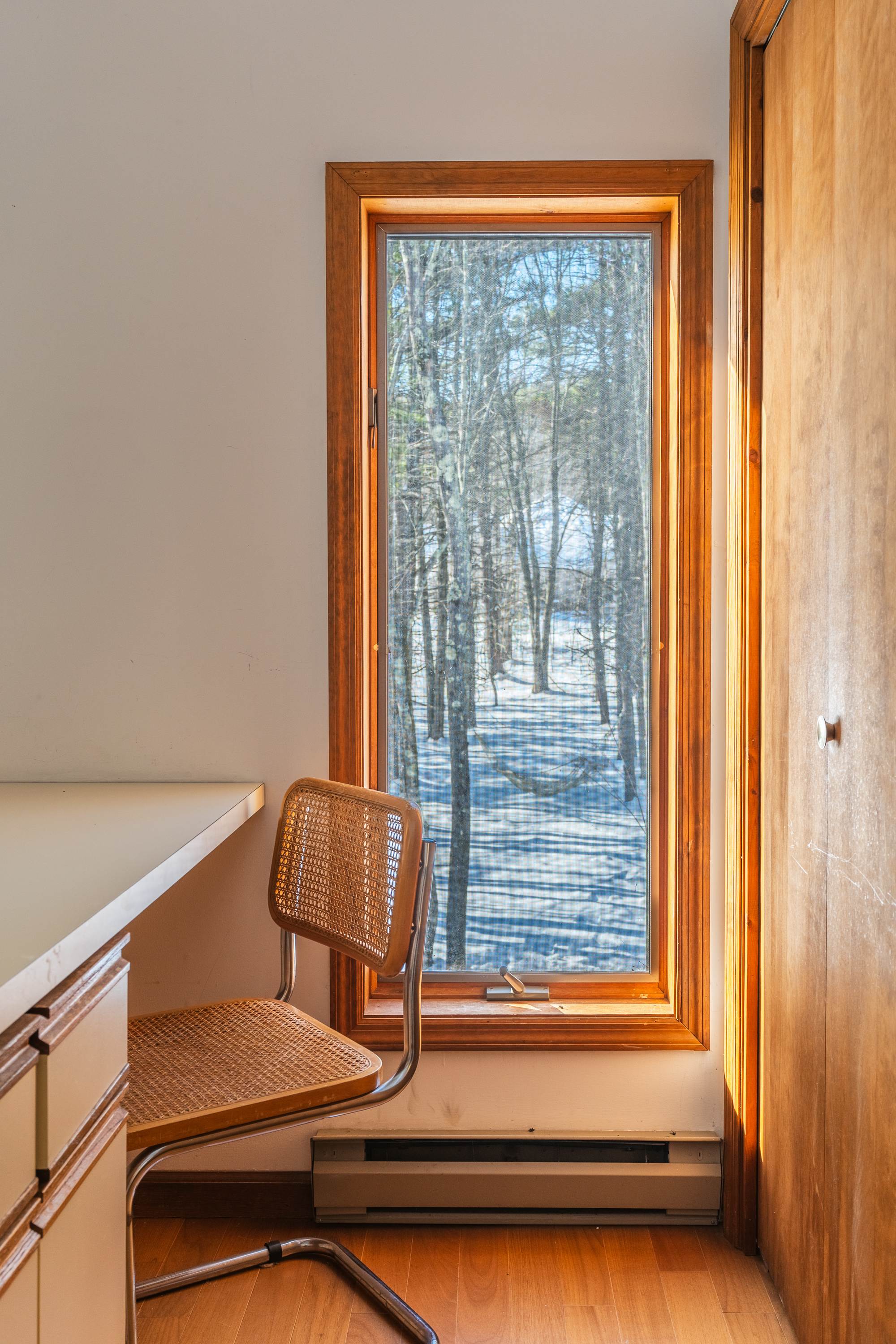 Small office nook with a built-in desk and a rattan chair, featuring hardwood floors. A large window provides natural light and a view of the snowy outdoor landscape.