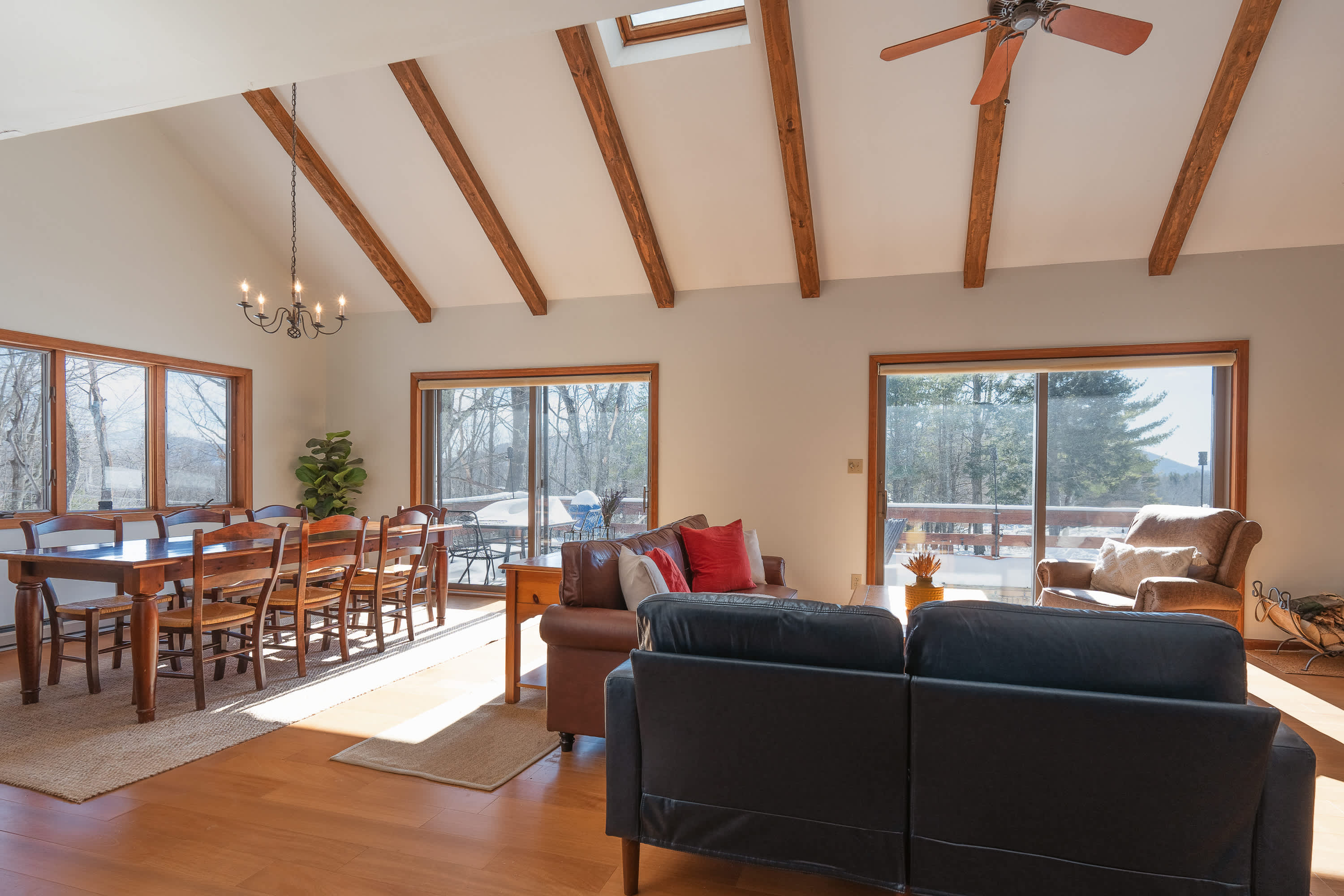 A bright, open-concept living and dining area features vaulted ceilings with exposed wooden beams, a blue sofa, and a brown leather couch, with large windows offering views of a snowy landscape.
