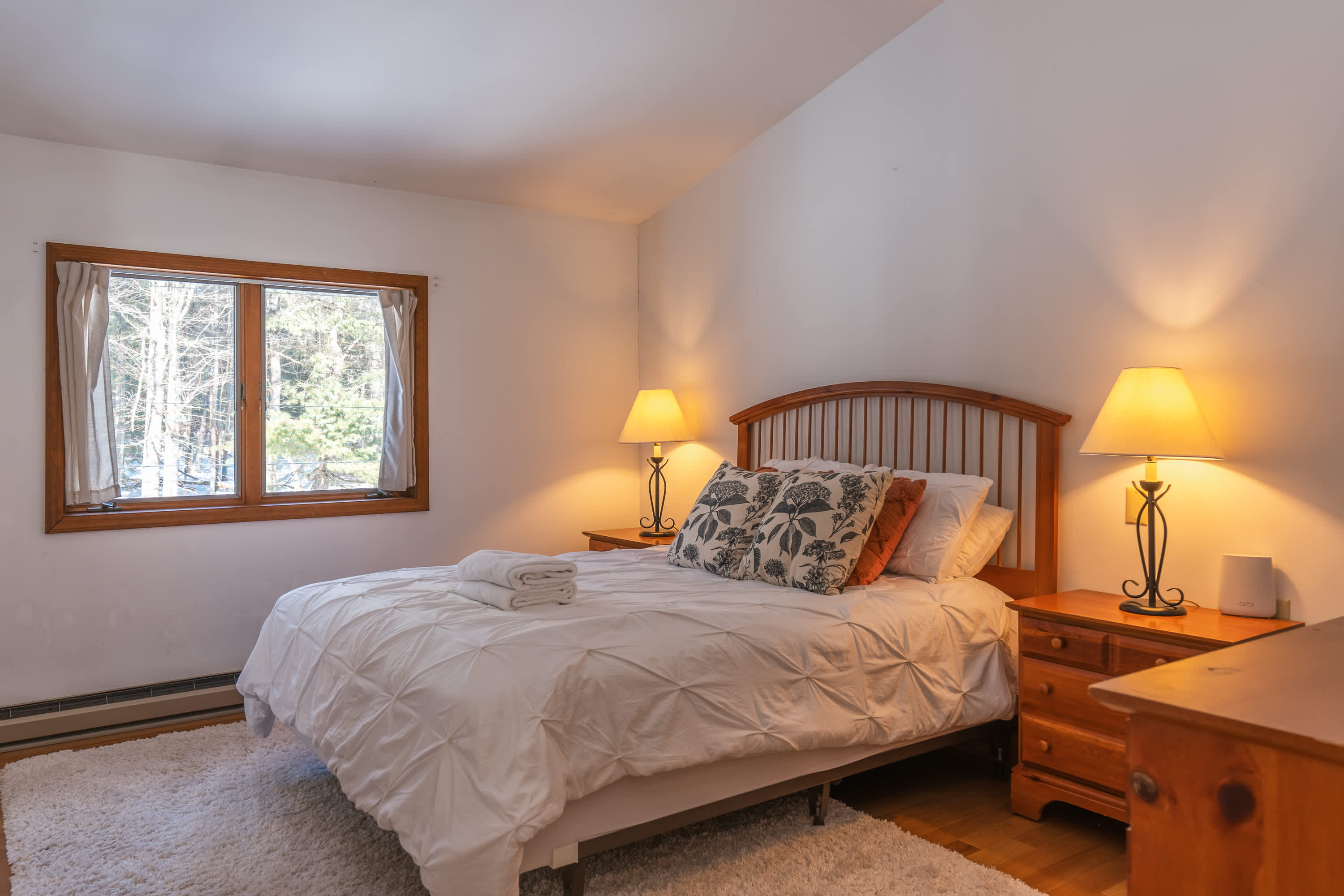 Cozy bedroom with a queen-sized bed, white bedding, and two bedside tables with lamps. Features hardwood floors, a plush white rug, and a window offering views of a snowy forest.