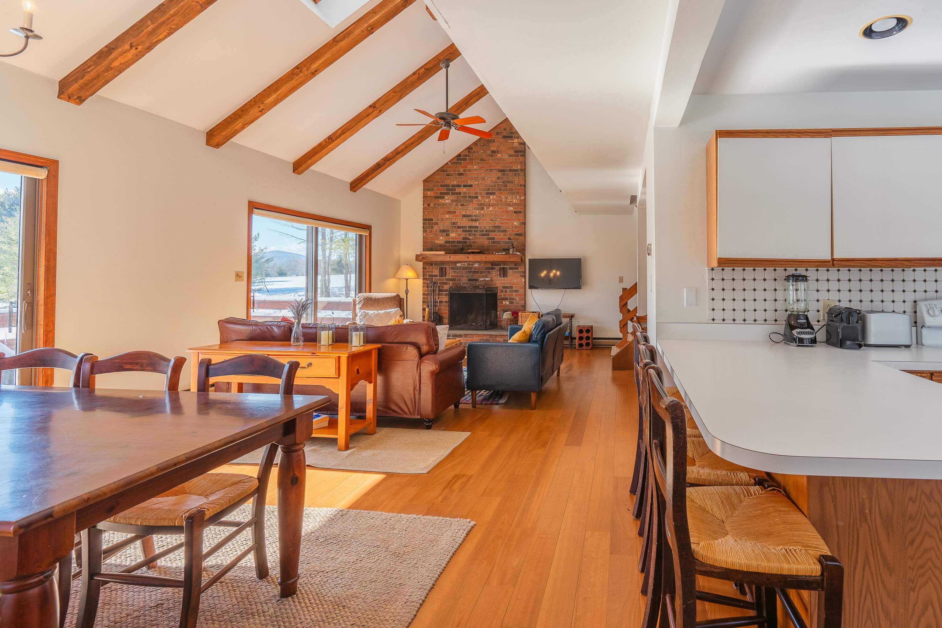 Spacious open-concept living area with vaulted ceilings, exposed wooden beams, and a large brick fireplace. Features comfortable leather sofas, a dining table, and large windows with snowy mountain views.