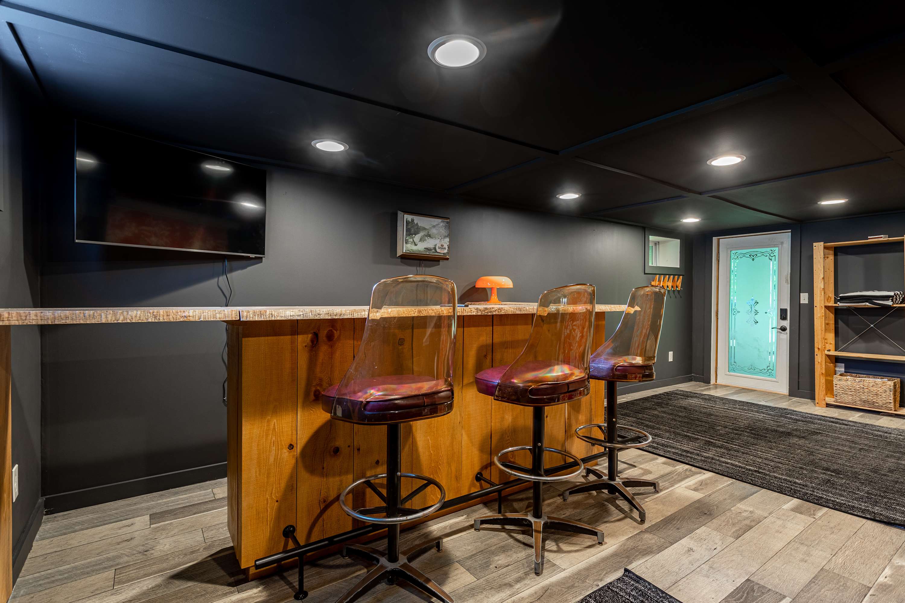 Another angle of the basement bar area, featuring a rustic wooden bar with three clear acrylic bar stools, recessed lighting, and dark gray walls. A glass-paned door leads to the exterior.