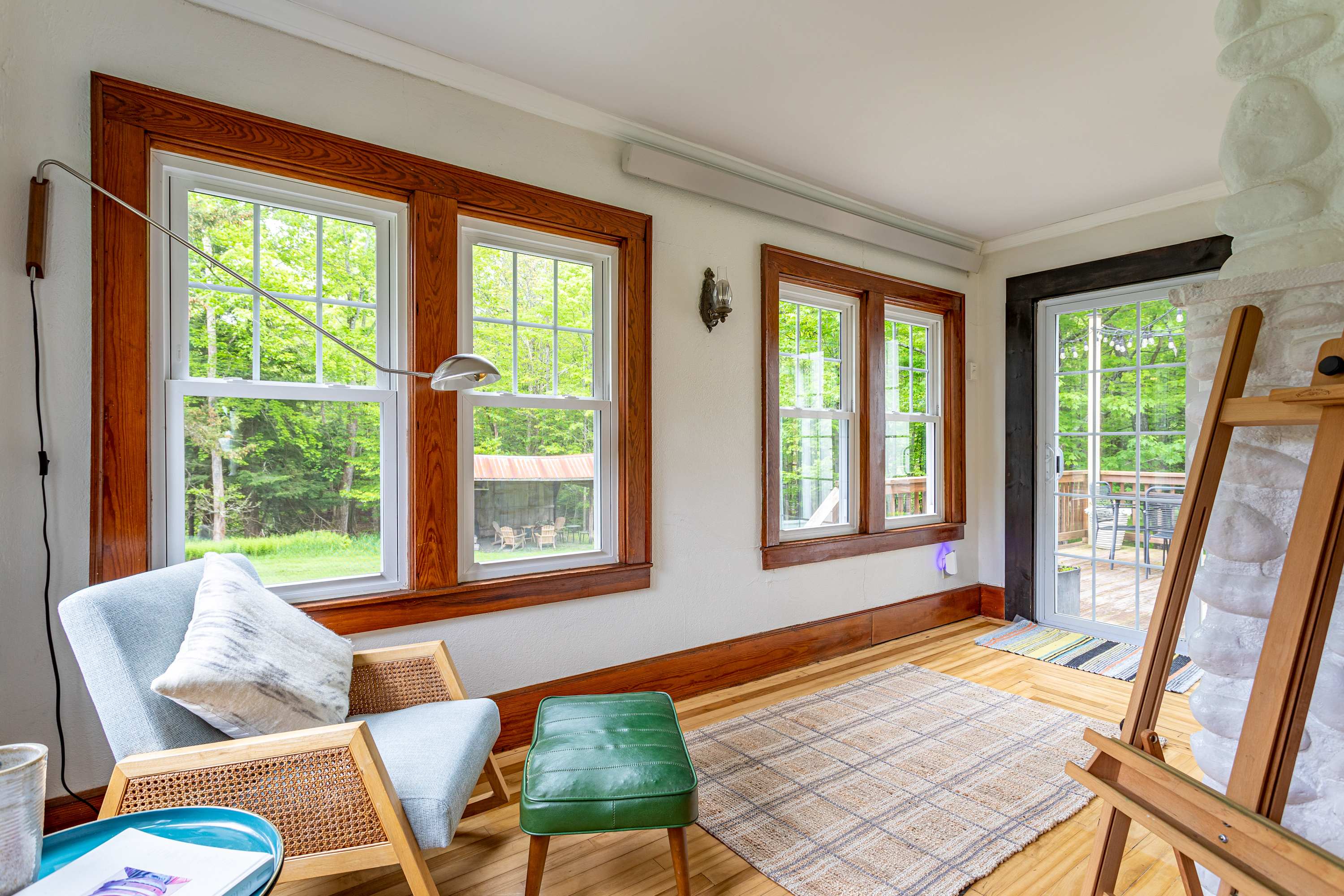 A bright sitting area with light hardwood floors and white walls, featuring multiple large windows with natural wood trim that offer expansive views of the lush green outdoors, furnished with a light blue armchair and a green ottoman.