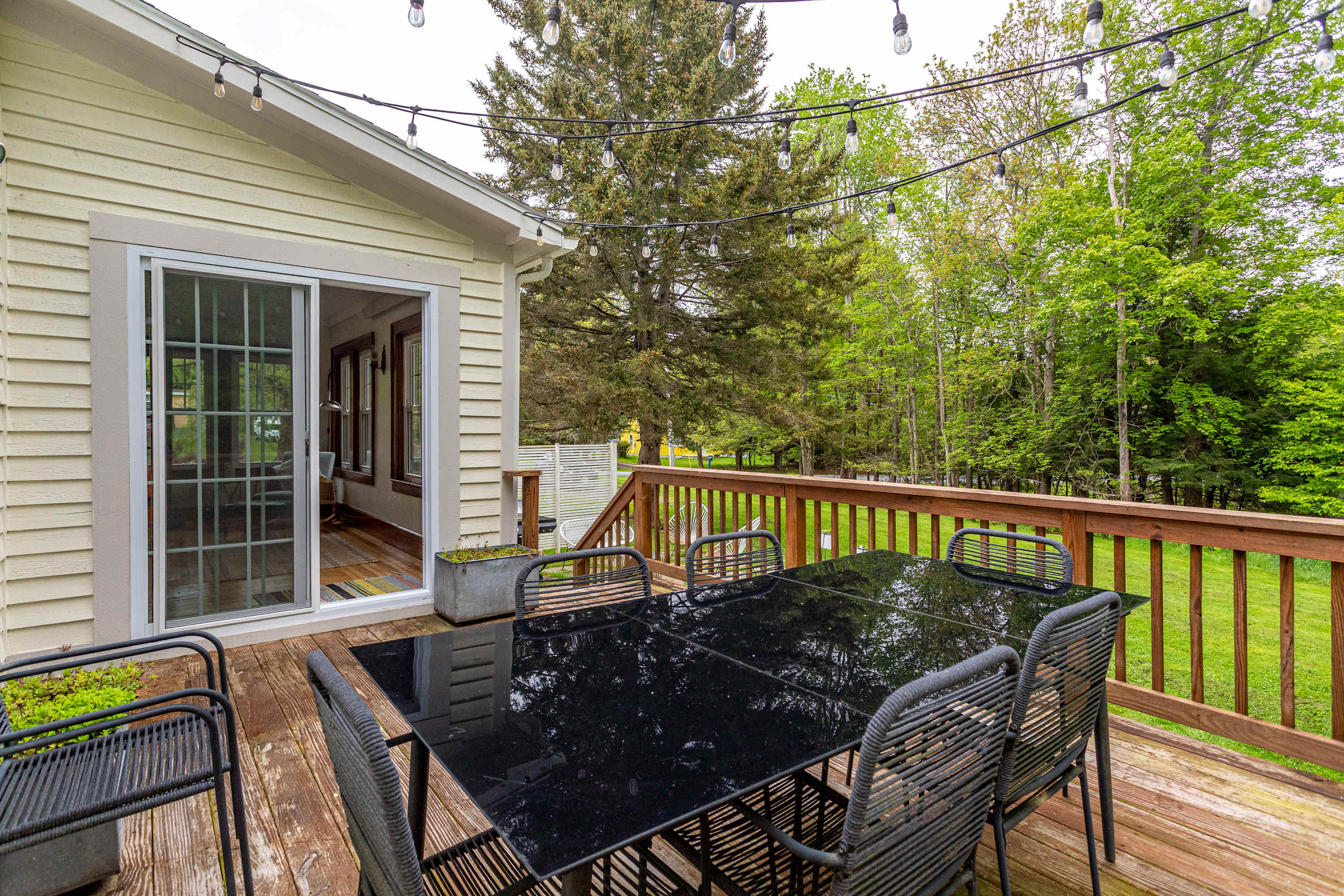 A spacious wooden deck features a black glass-top dining table with six chairs, string lights overhead, and a sliding glass door leading into the house, with lush green trees in the background.