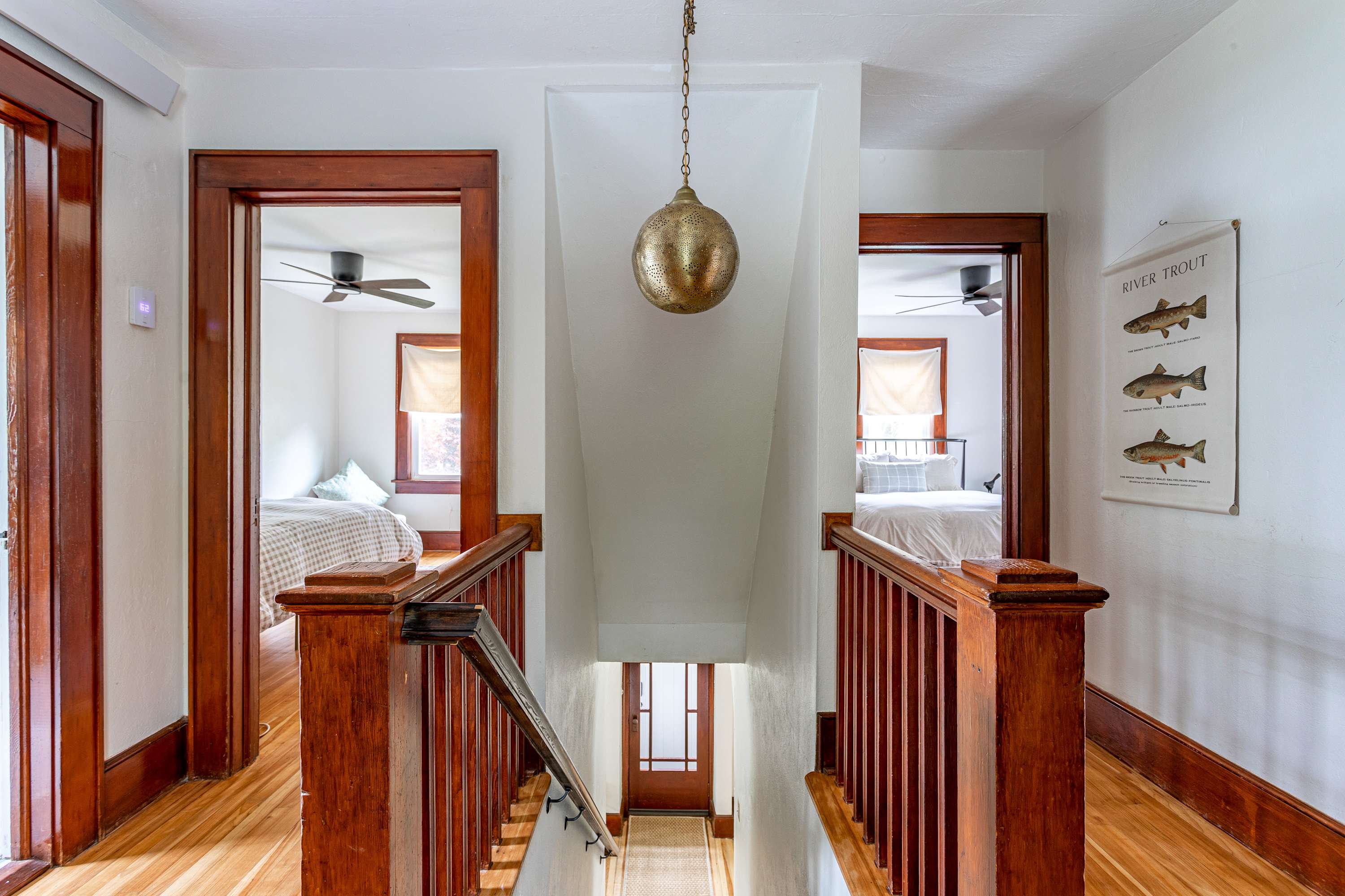 A bright upper-level foyer features a central staircase with dark wood railings, leading to a lower level, and two open doorways revealing separate bedrooms with beds and ceiling fans. A brass pendant light hangs from the ceiling.