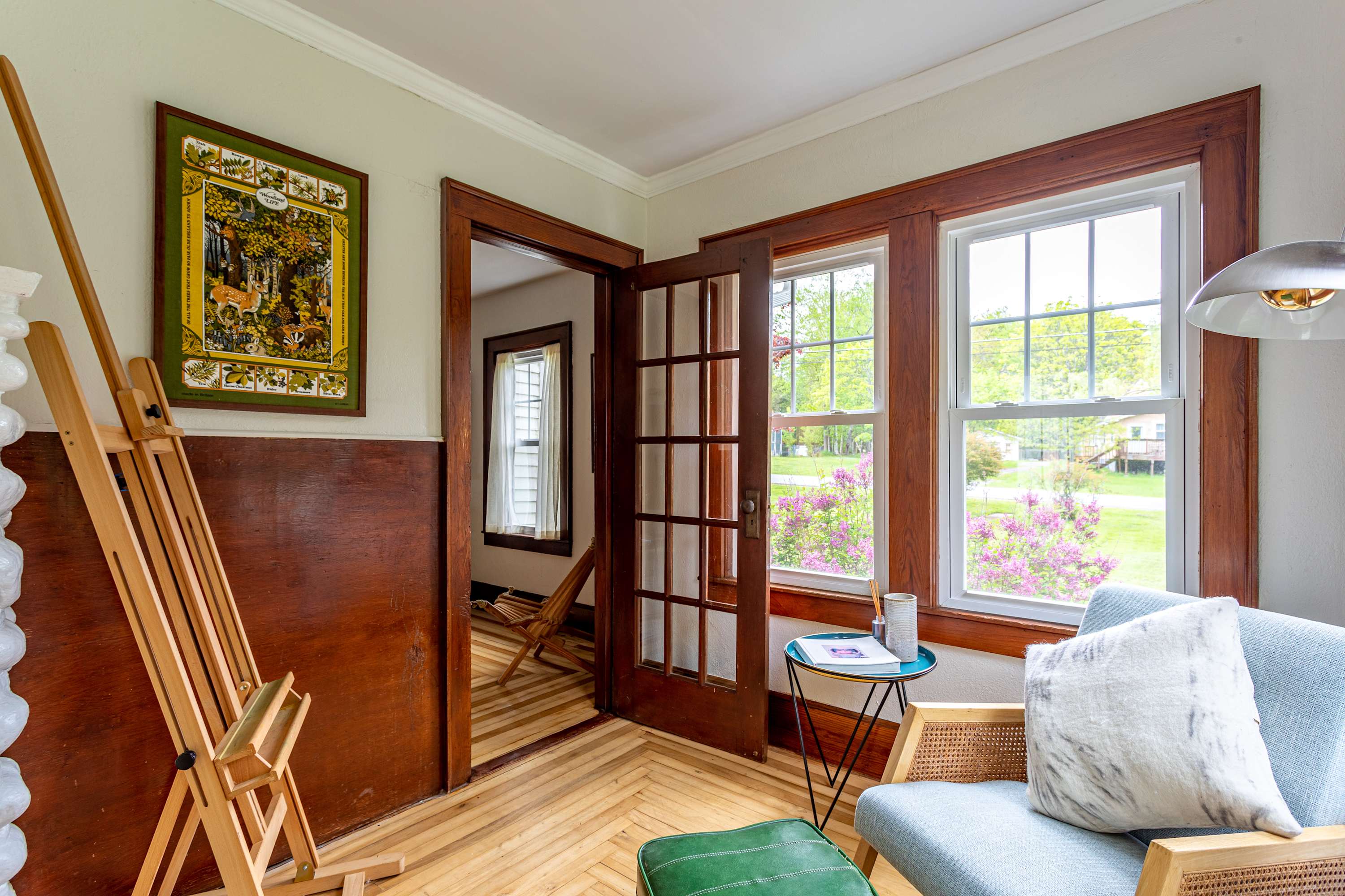 A bright, cozy office or reading nook with an easel, a light blue armchair, and large windows, featuring warm wooden paneling on one wall and hardwood floors.