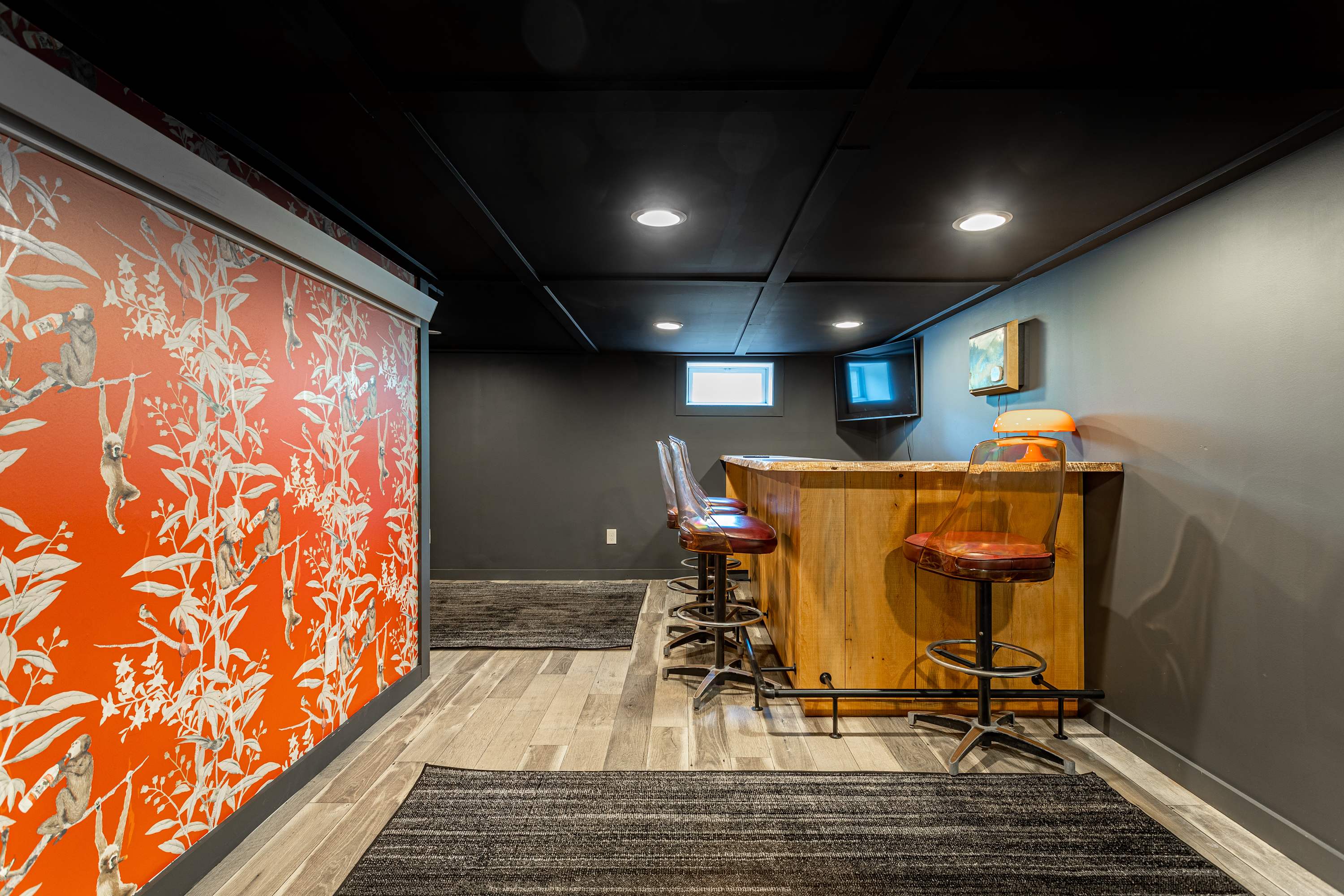 Stylish basement bar area with a rustic wooden bar, dark gray walls and ceiling, recessed lighting, and two vintage-style bar stools. A vibrant red and white patterned wallpaper accent wall adds a pop of color.