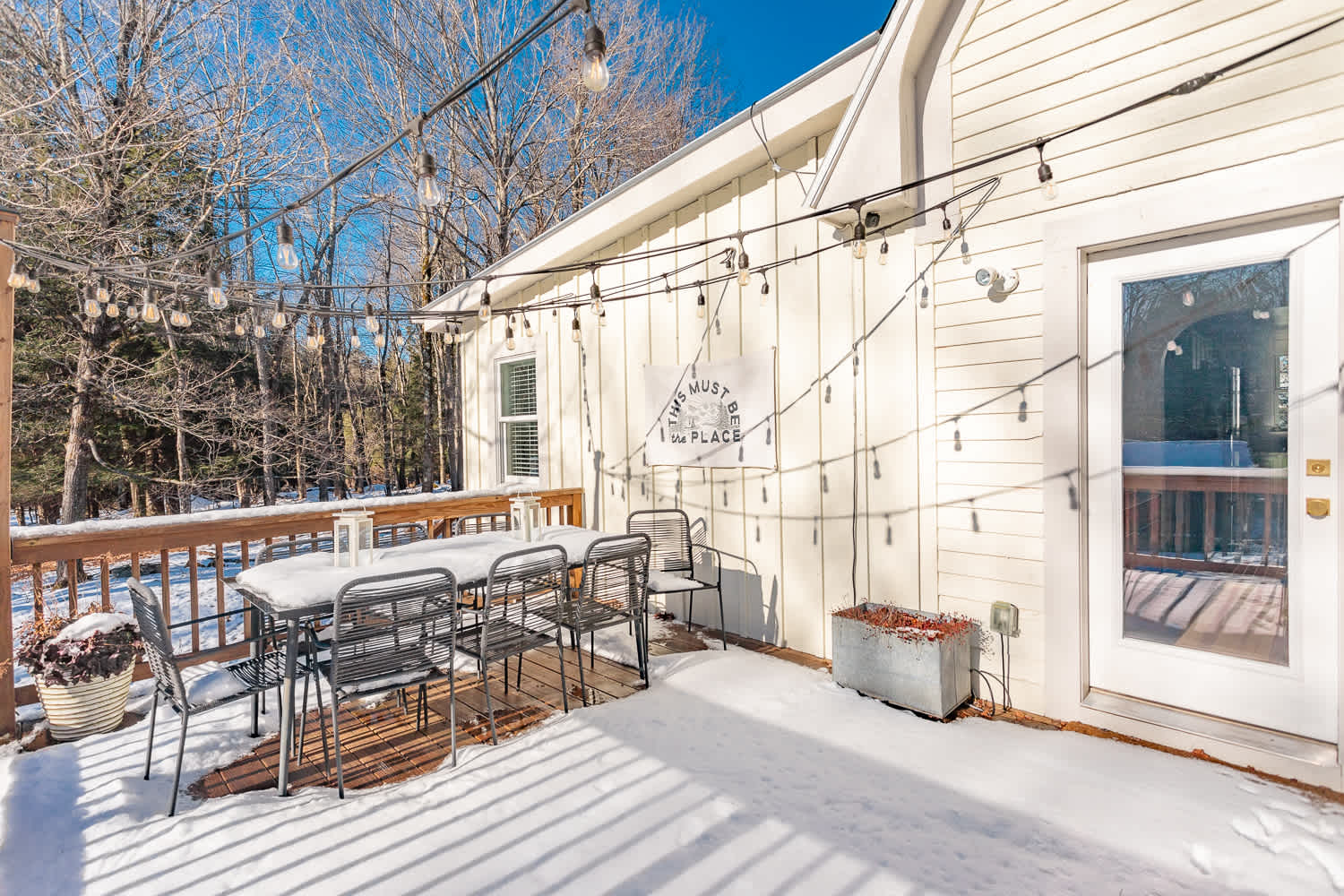A snowy wooden deck features a dark metal outdoor dining set with six chairs, illuminated by string lights, against a light-colored house with a "This Must Be The Place" sign. Bare trees line the background under a clear blue sky.