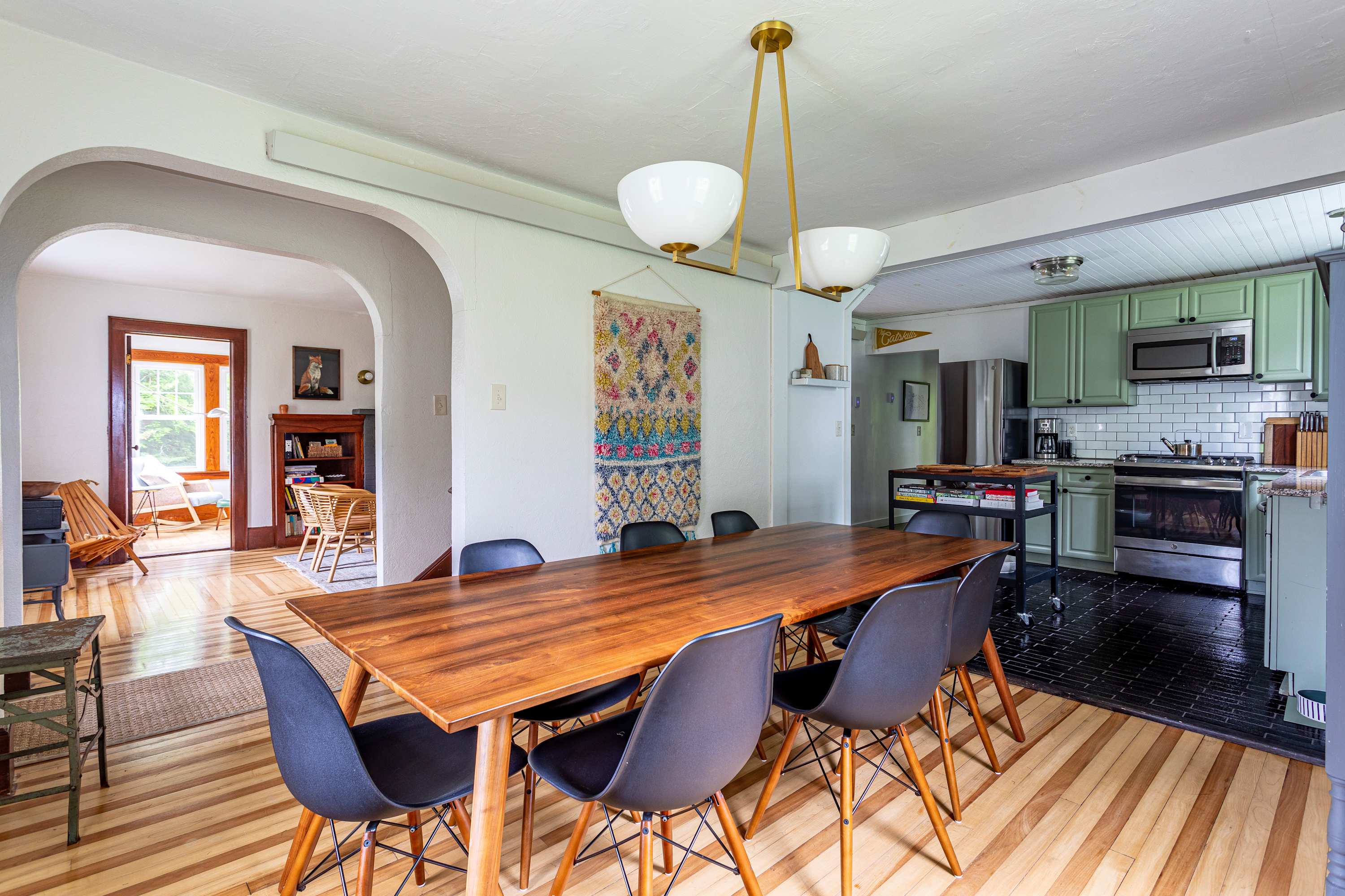 An open-concept dining area with a long wooden table and black chairs, seamlessly flowing into a kitchen featuring light green cabinets, black tiled floors, and modern stainless steel appliances.