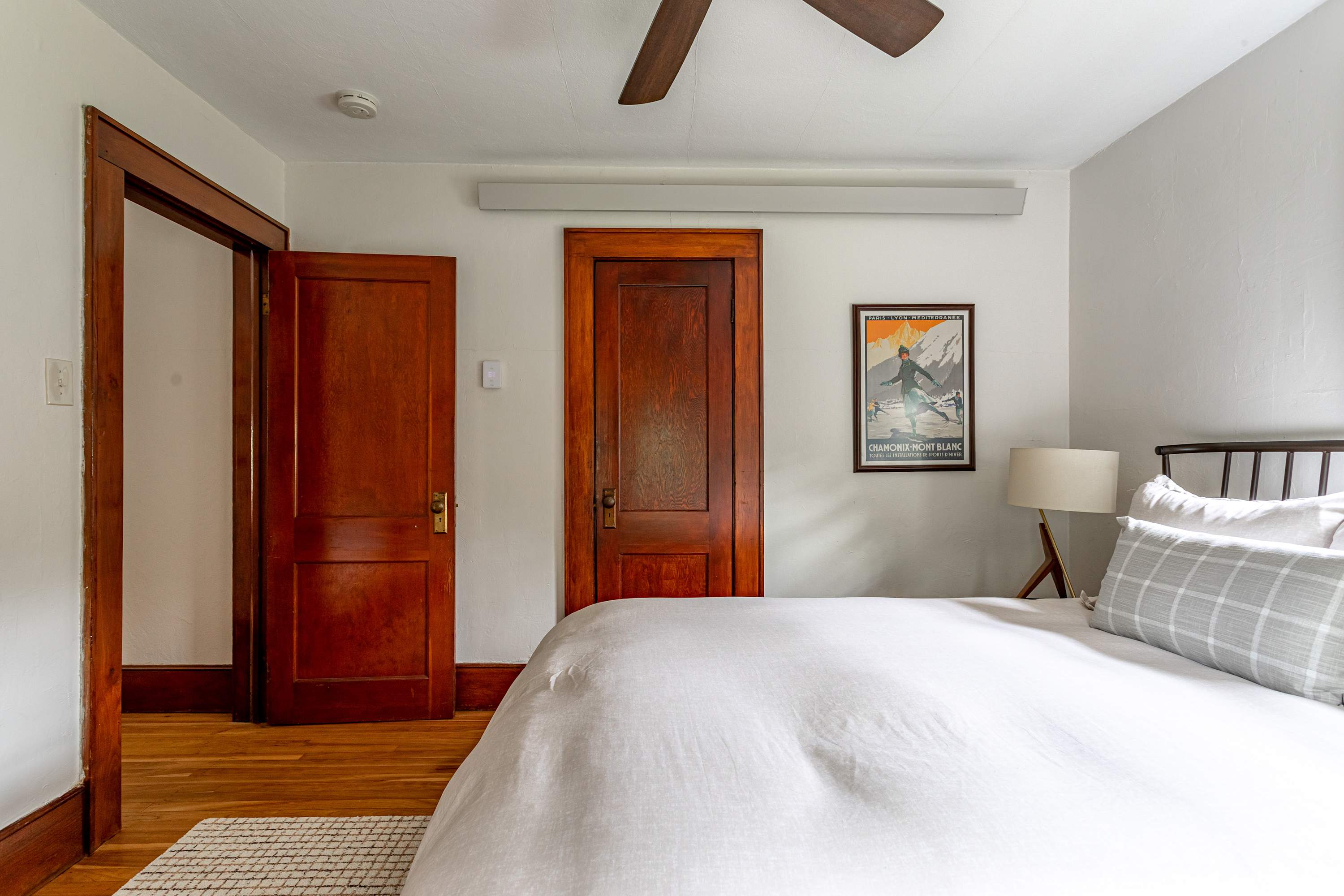 View of a bedroom with a black metal bed frame and white bedding, showing a closed wooden door leading to a closet and an open wooden door to the hallway. A vintage ski poster hangs on the wall.