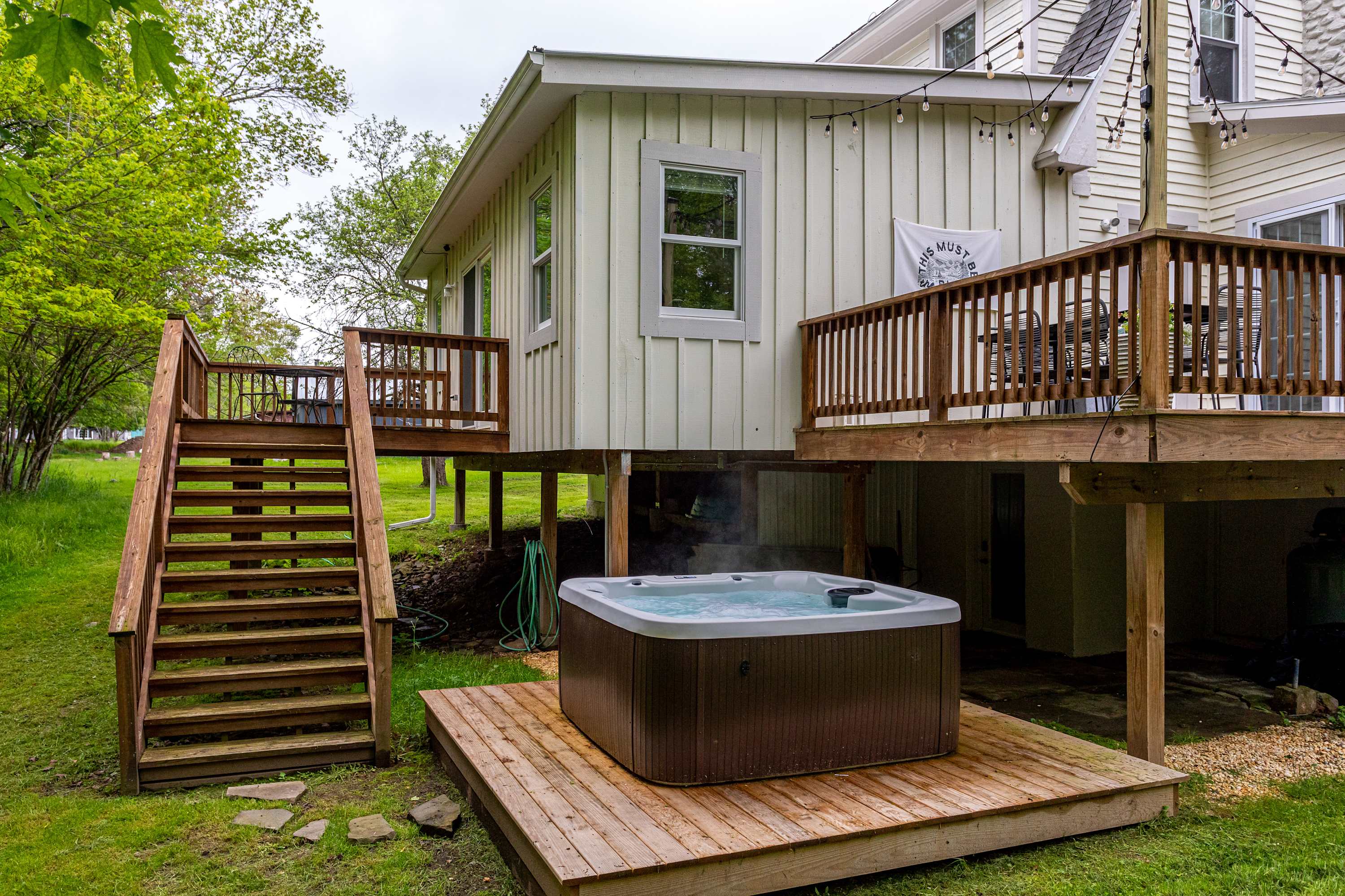 A hot tub on a wooden platform is positioned beneath a raised wooden deck with stairs leading up, set against the light-colored exterior of the house and surrounded by green foliage.