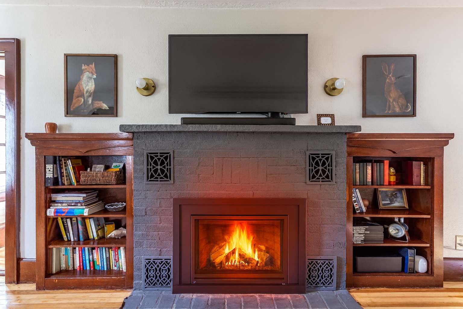 A cozy living room centered around a gray brick fireplace with a mounted television above, flanked by two built-in wooden bookshelves filled with books and decor, all set against light-colored walls.
