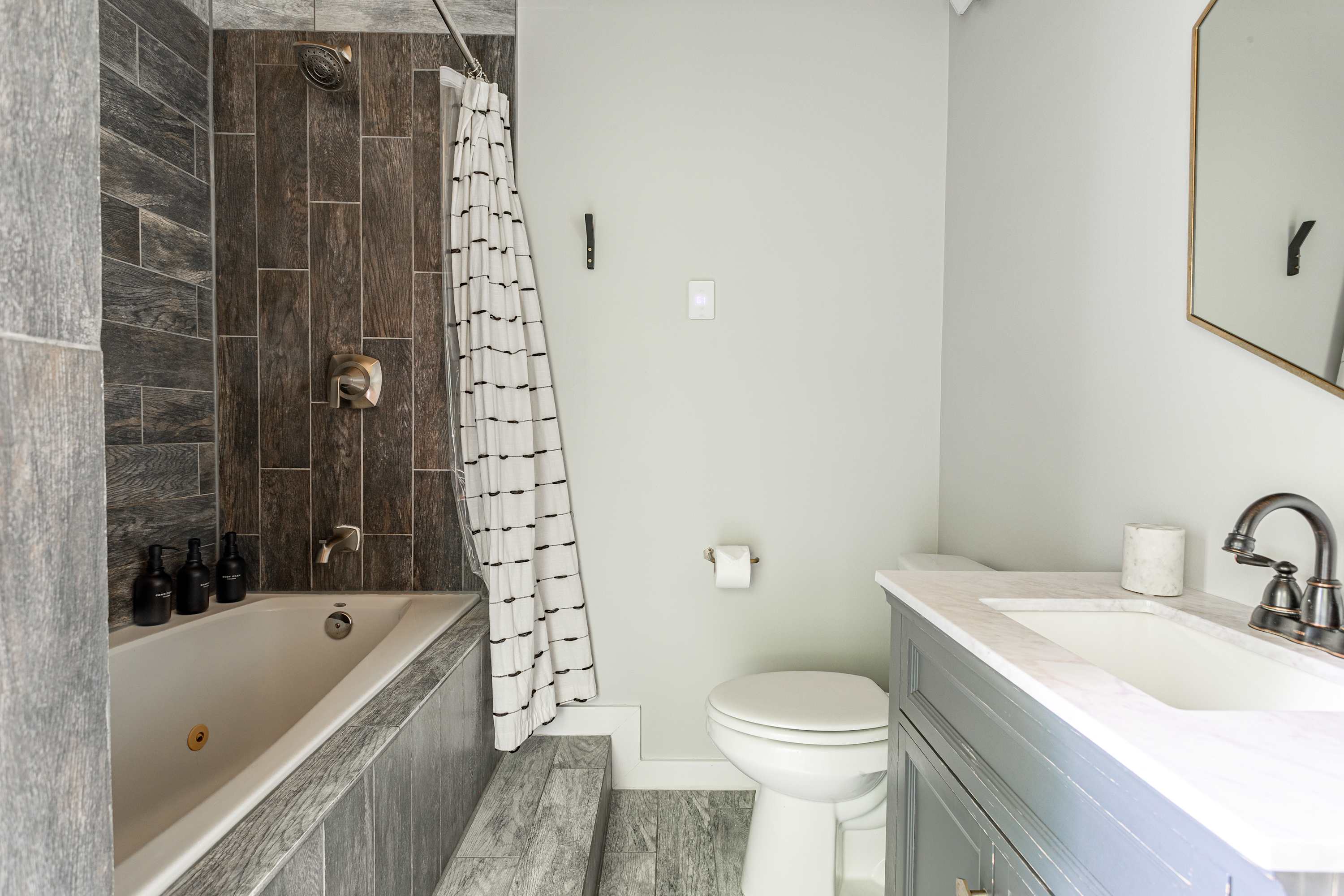 Modern bathroom with a deep soaking tub and shower featuring dark wood-look tile surround, a light gray vanity with a white sink, and a toilet. The walls are painted a light sage green, and the floor has gray wood-look tiles.