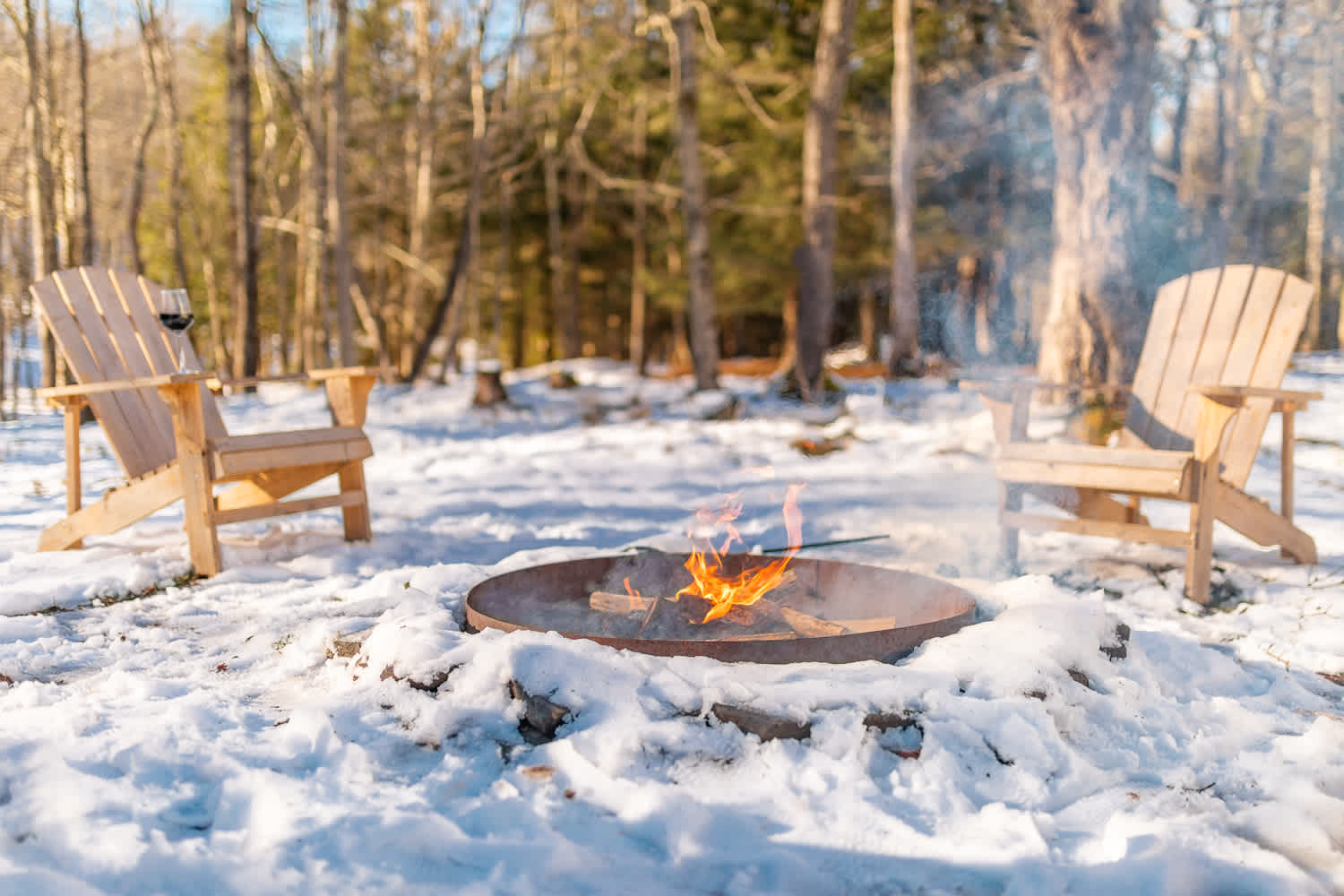 Two wooden Adirondack chairs flank a metal fire pit with a small fire burning, surrounded by a blanket of snow in a wooded area with bare trees.