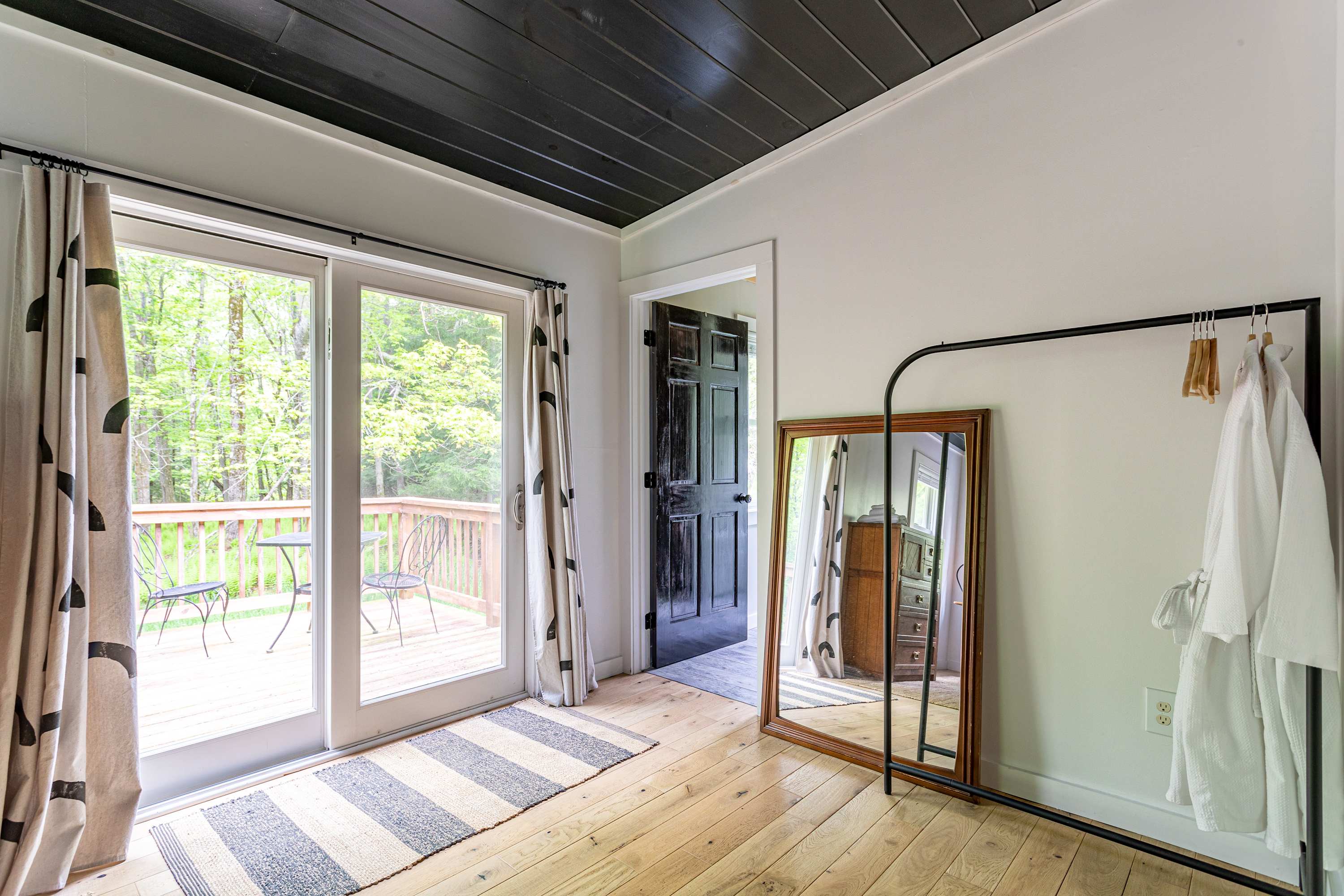 This bedroom offers sliding glass doors leading to an outdoor deck, featuring a full-length mirror, a clothing rack, and light hardwood floors, with a dark door visible in the background.