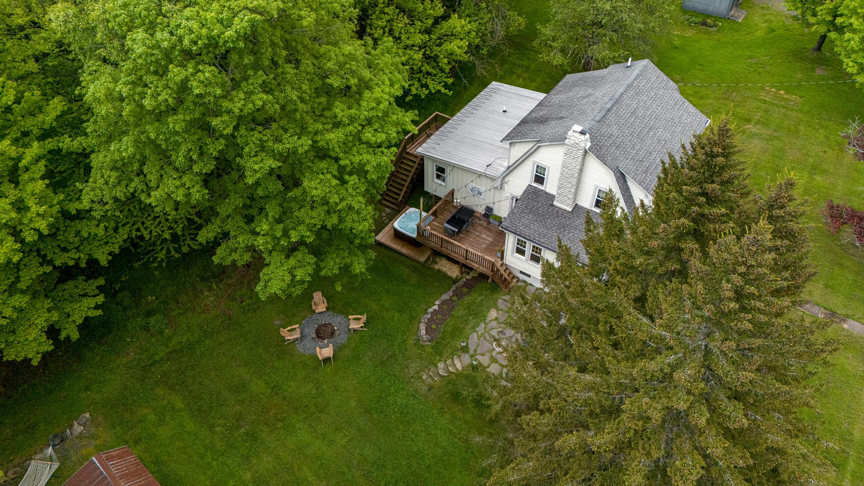 An aerial view shows a white house with a gray roof nestled among lush green trees and grass, featuring a wooden deck with a hot tub and a fire pit area with chairs in the yard.