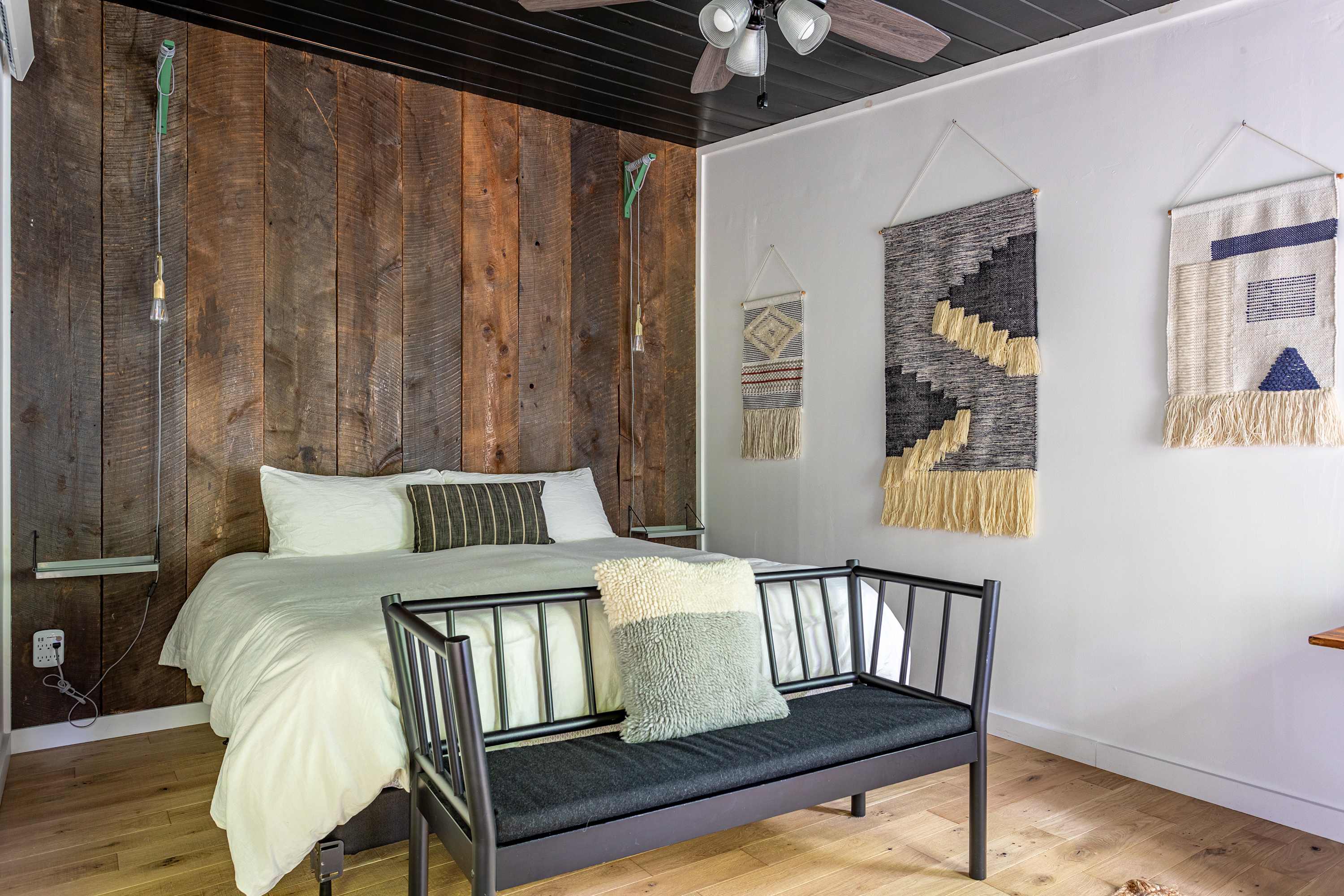 A stylish bedroom featuring a large bed against a rustic wooden accent wall, a black paneled ceiling, and decorative wall tapestries, with a black bench at the foot of the bed.