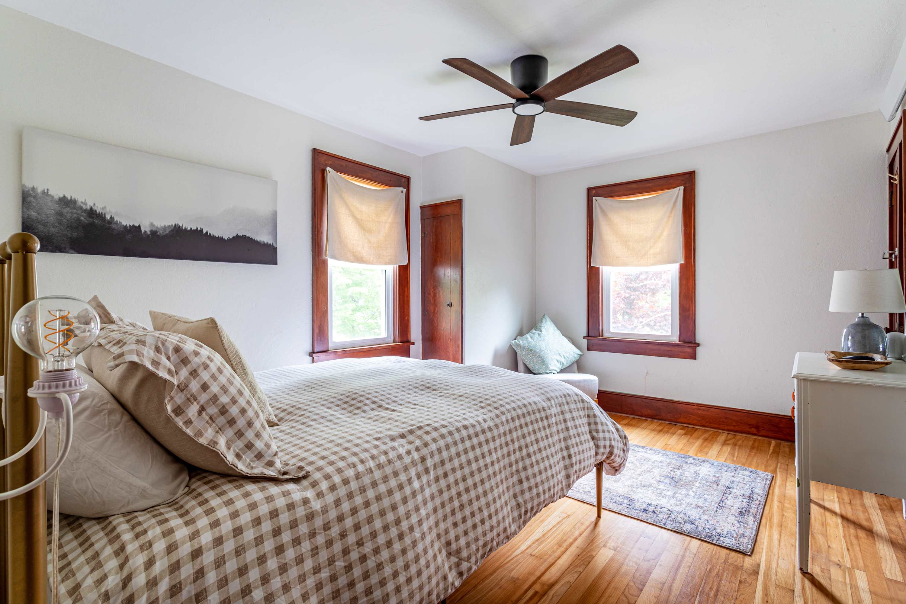 Cozy bedroom with a brass-colored metal bed frame, checkered duvet, two windows with natural light, a dark wood ceiling fan, and a white dresser with a lamp. Hardwood floors and white walls complete the look.