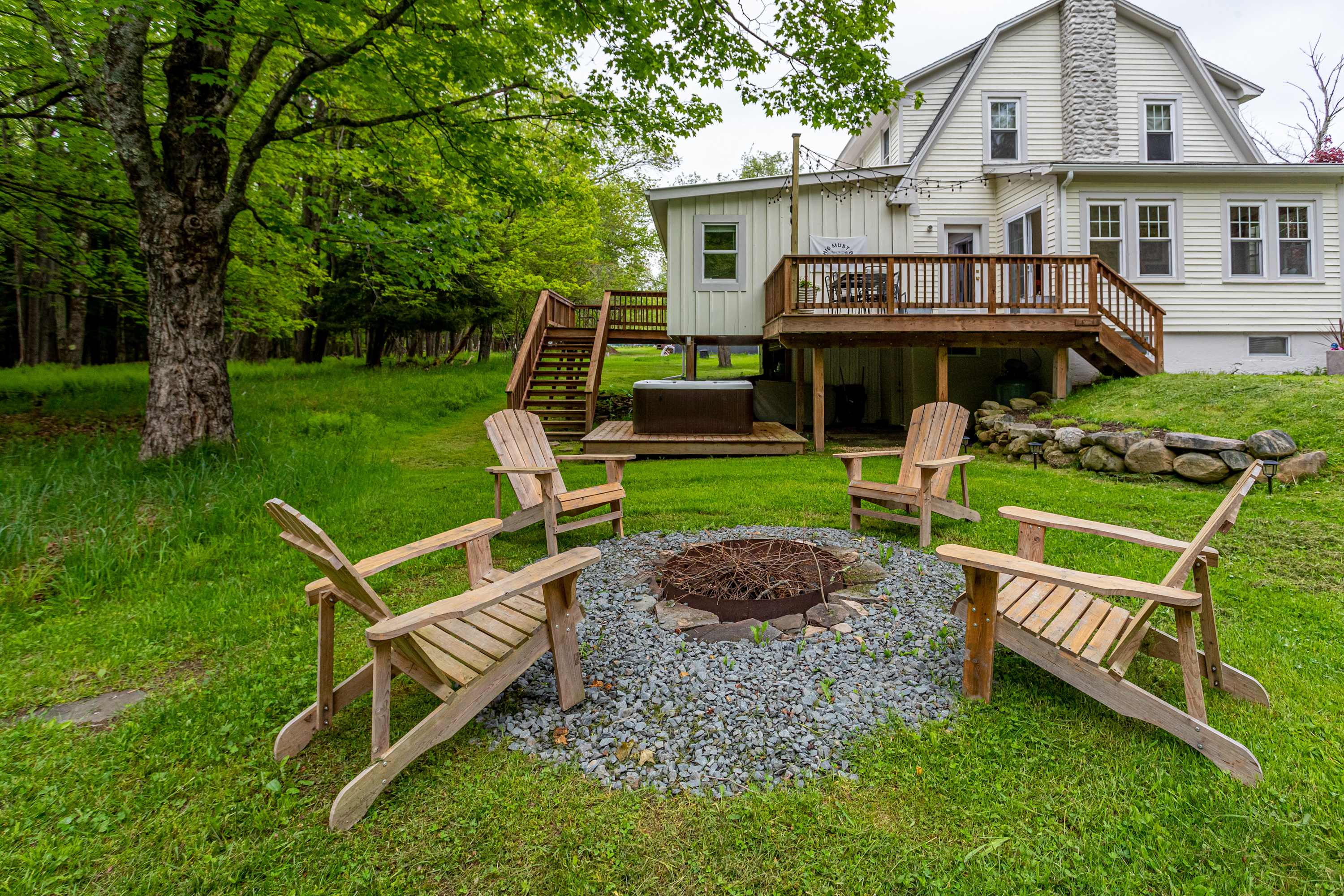The back exterior of the house features a wooden deck and a lush green yard with a circular stone fire pit surrounded by four wooden Adirondack chairs, with a hot tub visible below the deck.