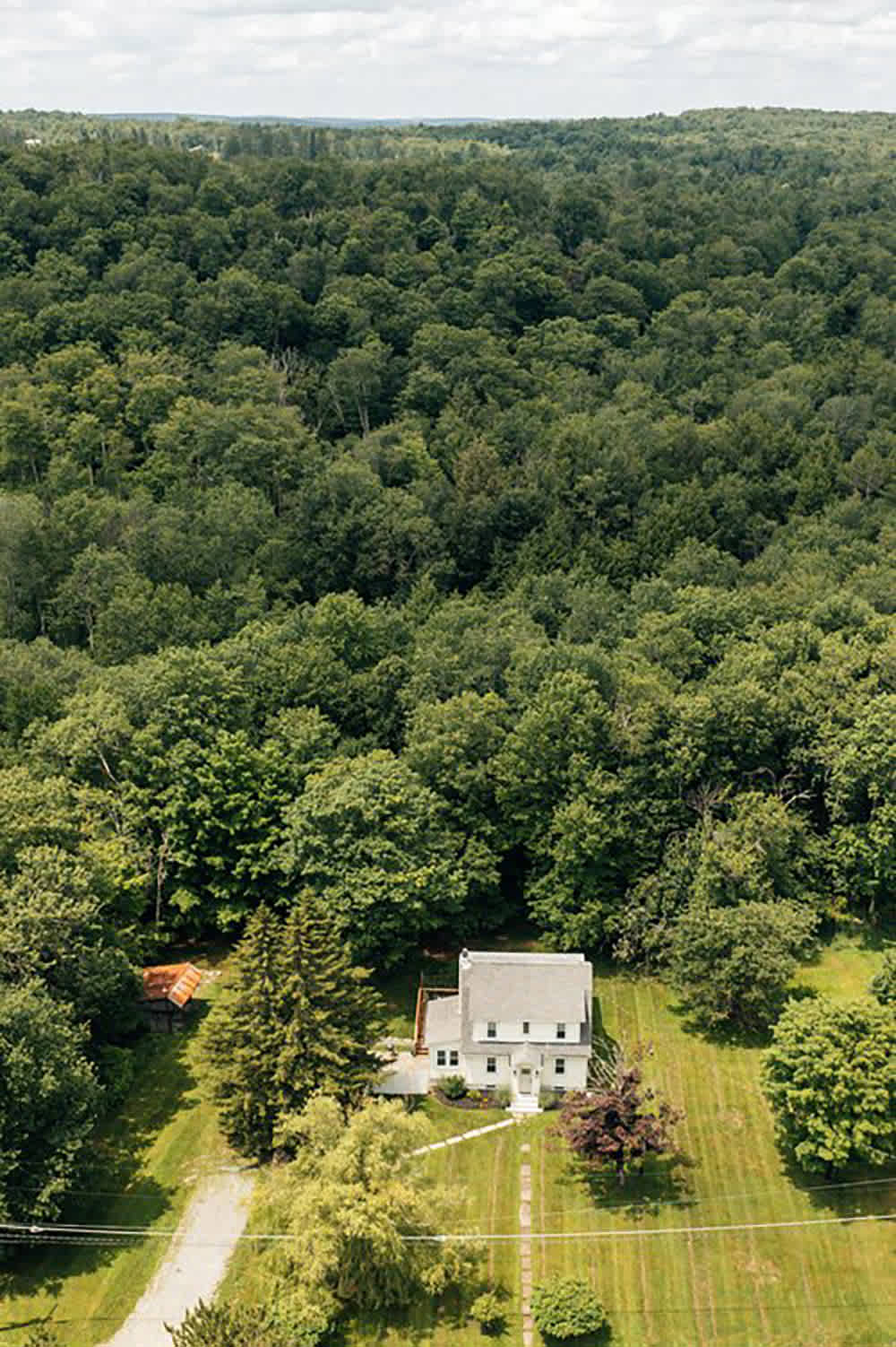 An aerial view reveals a white house with a gray roof situated in a clearing, surrounded by vast, dense green forests under a bright, partly cloudy sky, highlighting the property's secluded natural setting.
