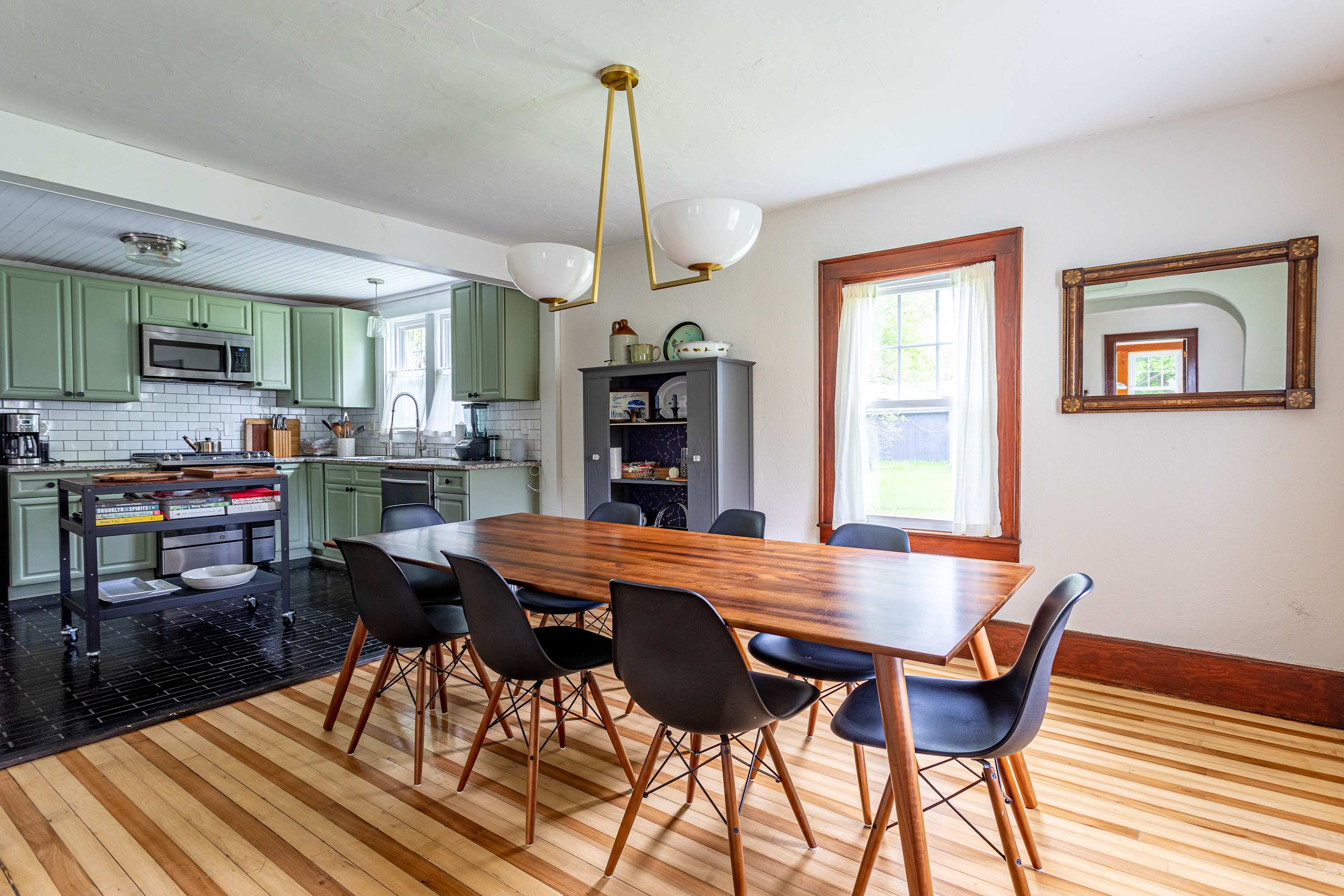 The dining room features a large wooden table and black chairs, with a view into the kitchen showcasing light green cabinets and a black tiled floor, under a modern brass chandelier.