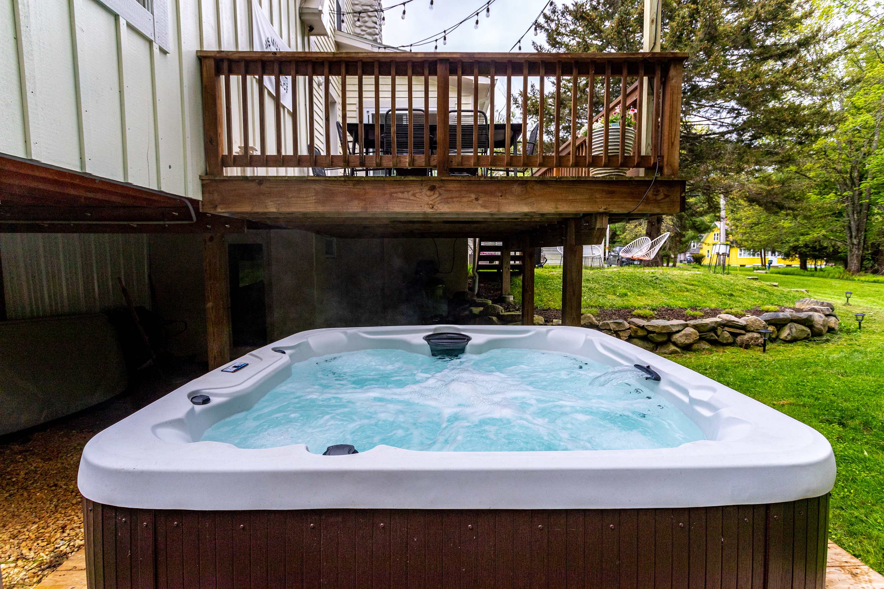 A bubbling hot tub is nestled beneath a raised wooden deck, adorned with string lights, offering a private and relaxing outdoor space adjacent to the white siding of the house and a grassy area.