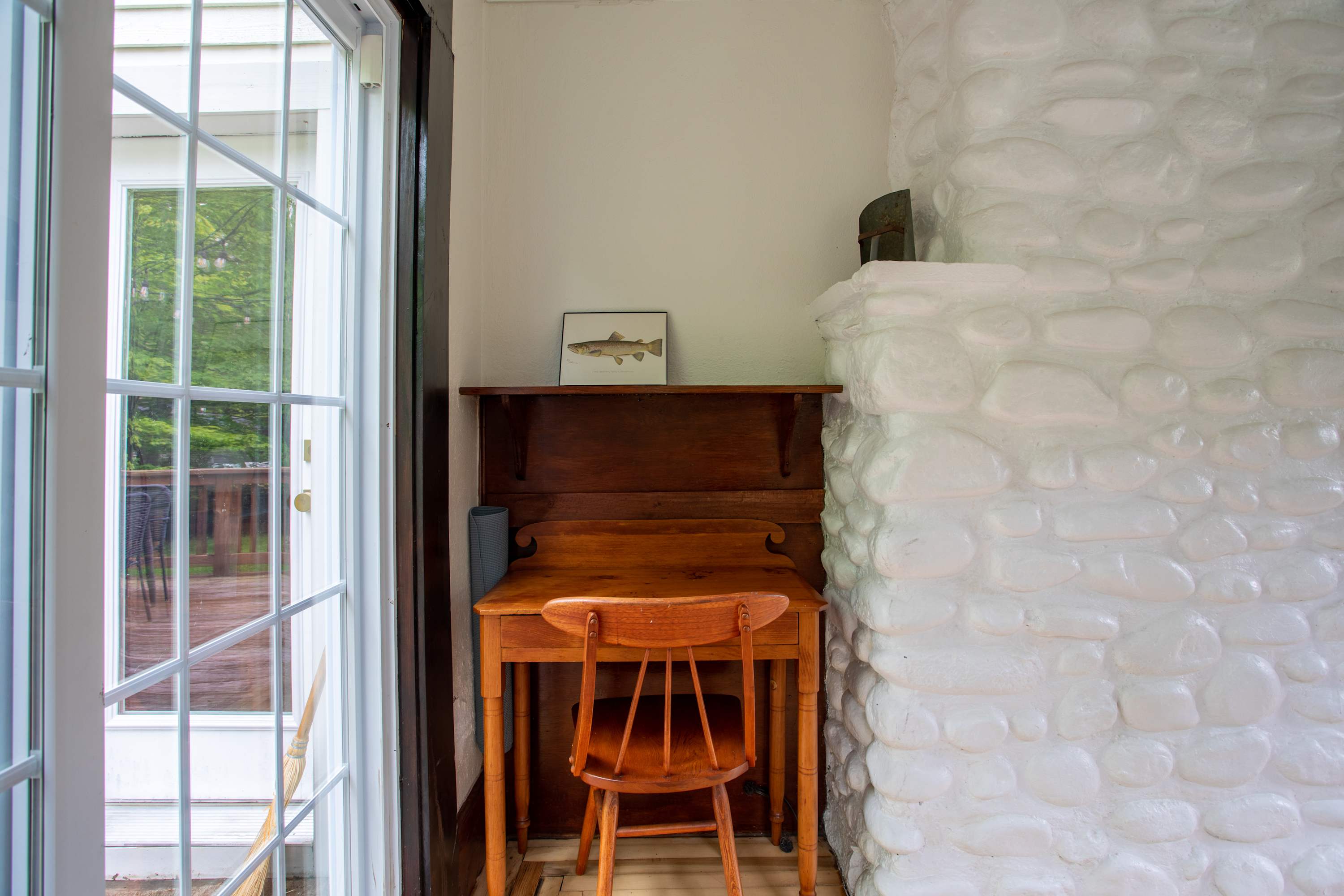 A compact office space featuring a classic wooden desk and chair, positioned next to a textured white stone fireplace, with a glass door providing outdoor access.
