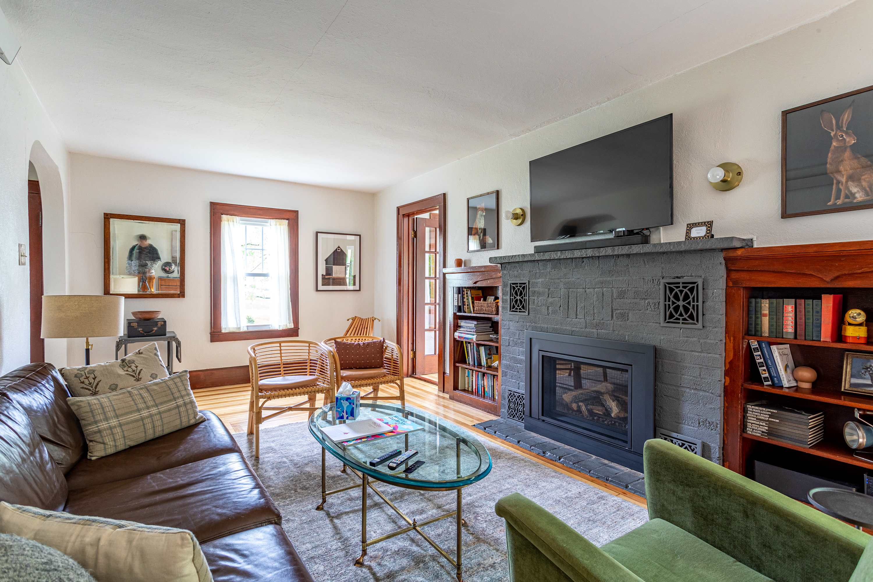 A cozy living room featuring a brown leather sofa, a glass coffee table, and a dark gray brick fireplace with a mounted TV, complemented by hardwood floors and rattan chairs.