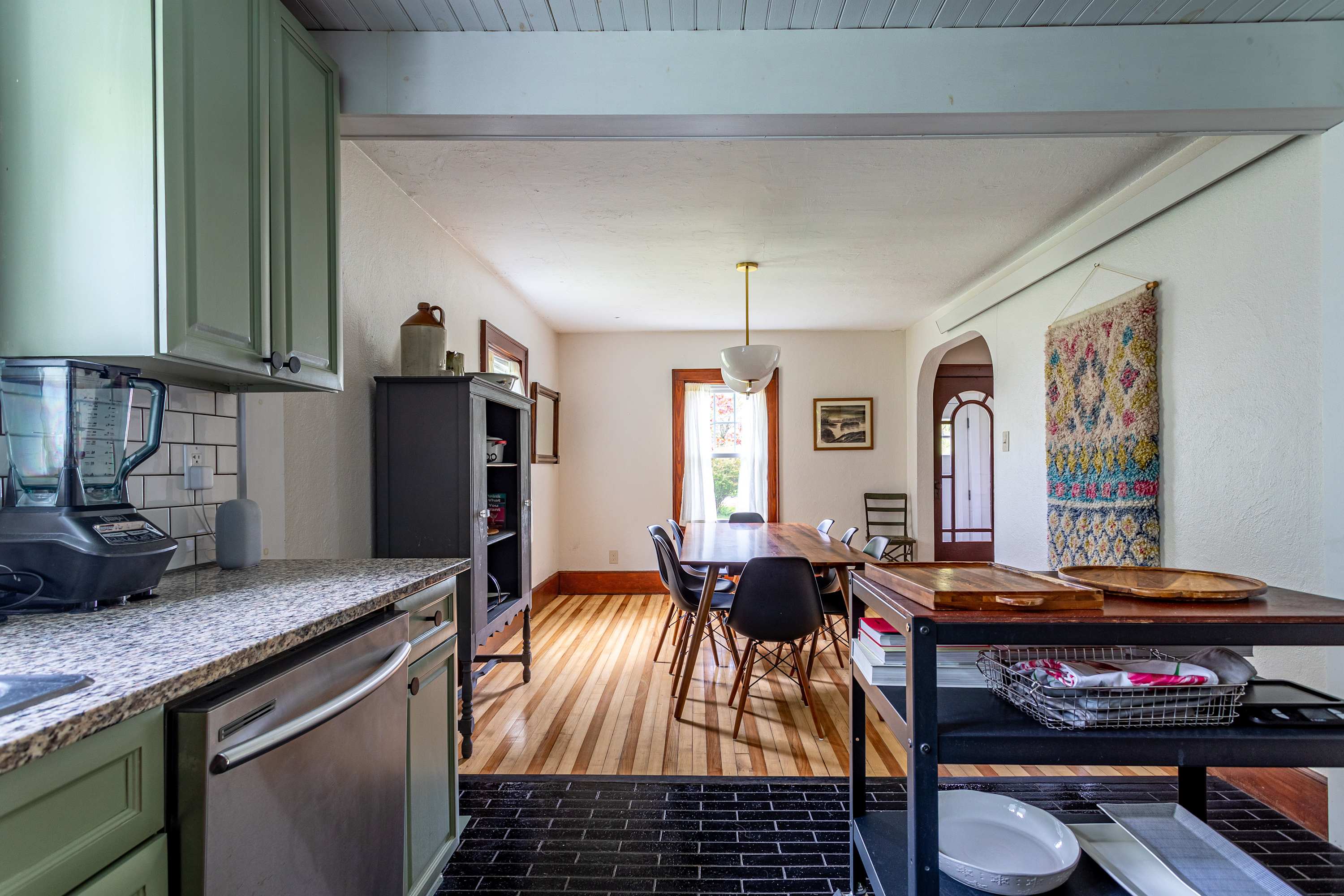 A bright kitchen with light green cabinets, granite countertops, and a white subway tile backsplash, opening into a dining room with a long wooden table and black chairs.
