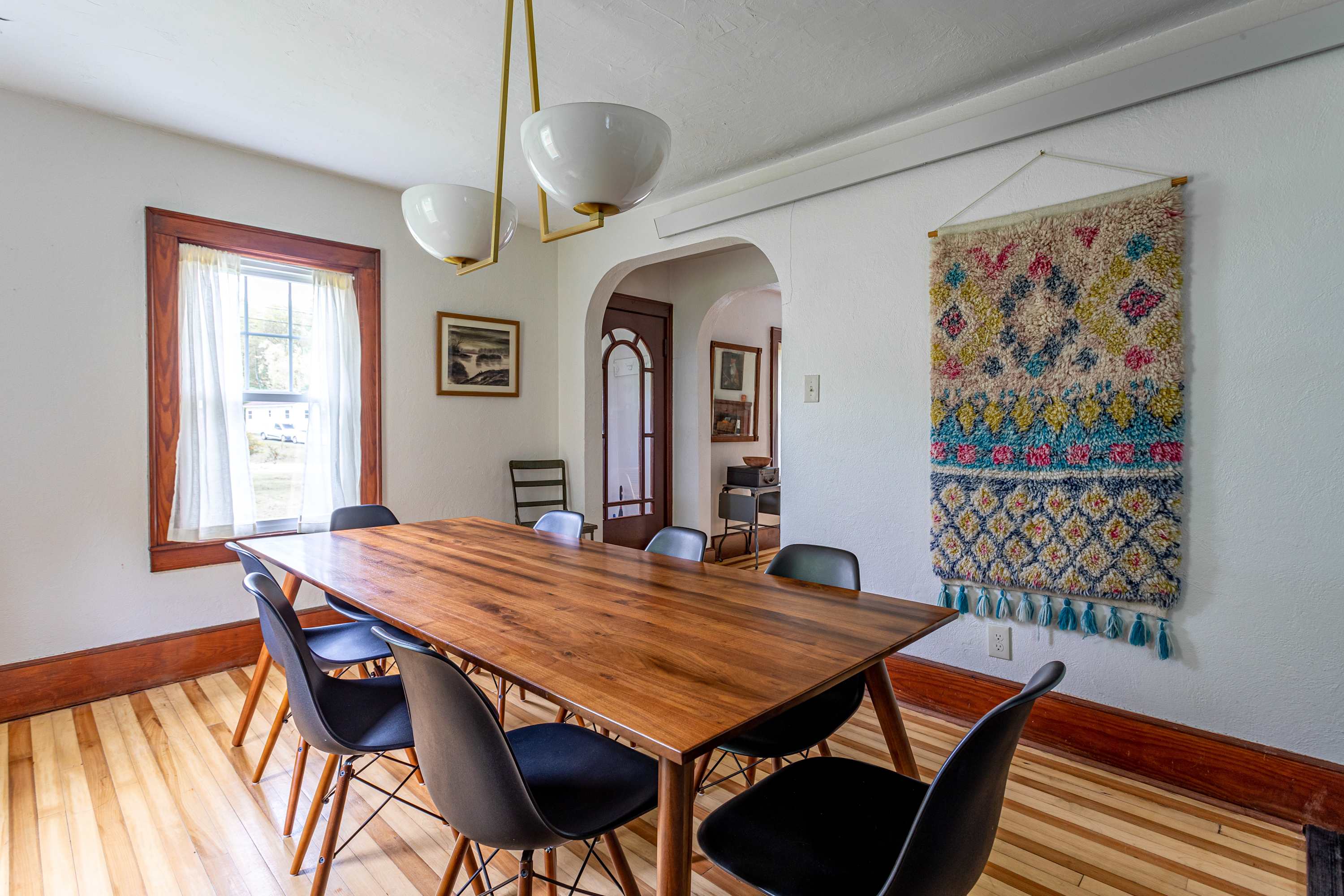 A spacious dining room featuring a long wooden table with black modern chairs, illuminated by a stylish brass and white chandelier, with hardwood floors and a colorful wall tapestry.