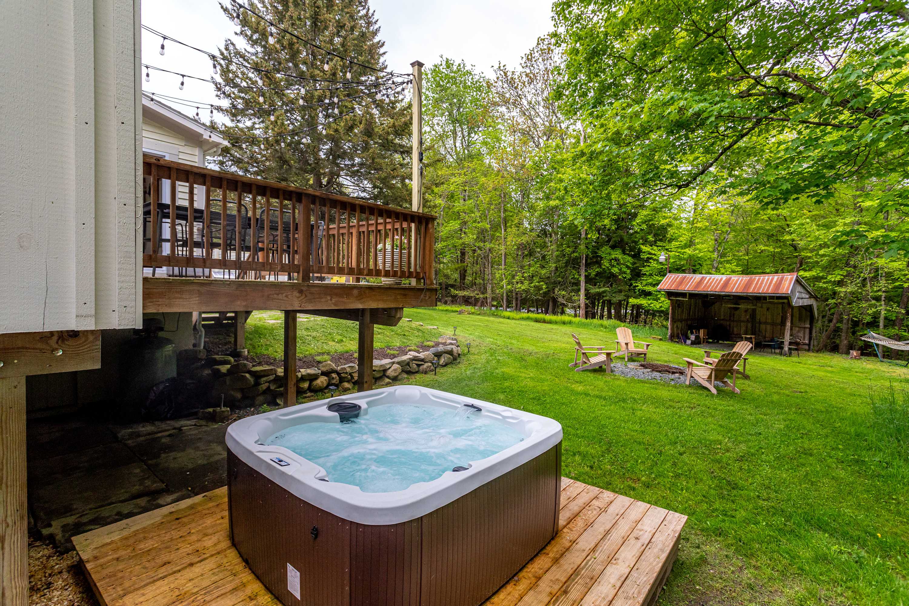 A bubbling hot tub sits on a wooden platform below a raised wooden deck, overlooking a lush green lawn with a fire pit area and a rustic shed in the background.