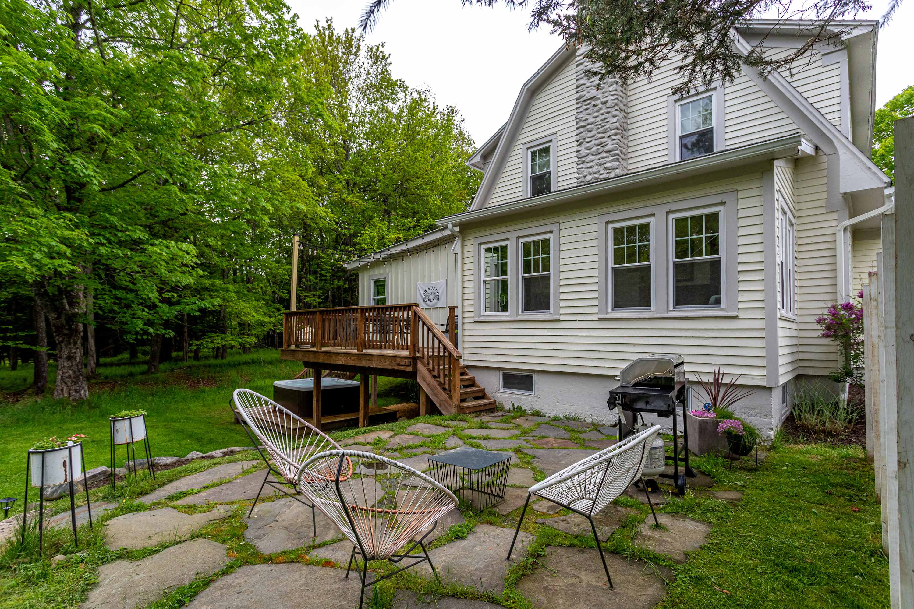 The back of a light-colored house with a stone chimney features multiple windows and a wooden deck, with a stone patio area and a BBQ grill on a green lawn surrounded by trees.