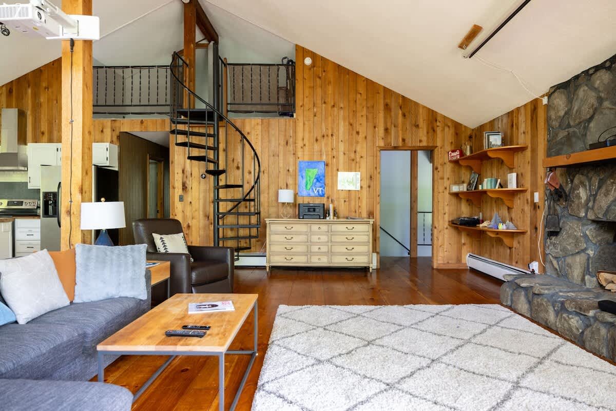 Spacious living room with rustic wood-paneled walls, a large stone fireplace, a gray sectional sofa, and a wooden coffee table. A black spiral staircase leads to a loft area, complementing the vaulted ceiling.