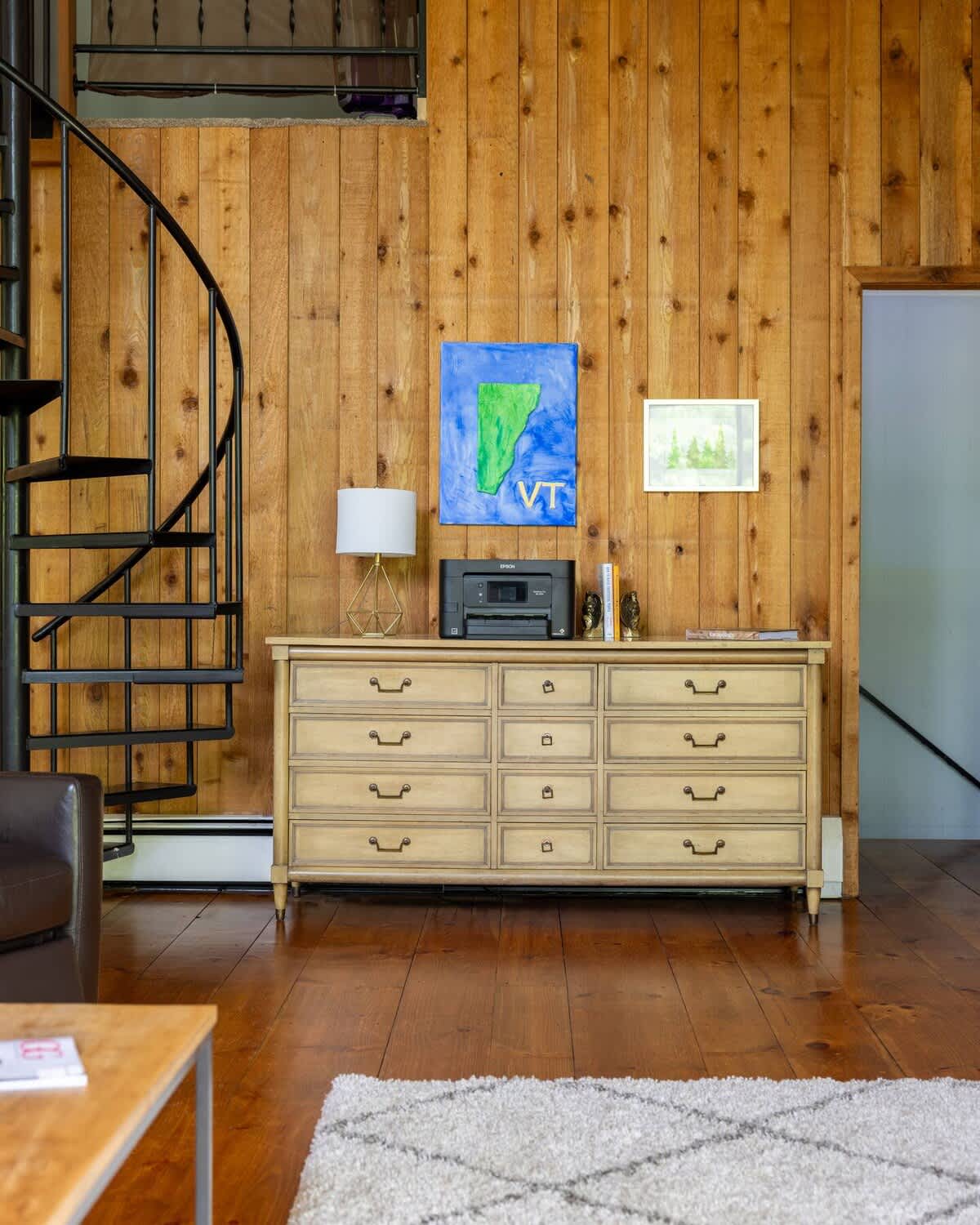 A corner of the living area featuring a black spiral staircase leading to an upper level, adjacent to a light-colored dresser with a printer and decorative artwork, against a warm wood-paneled wall.