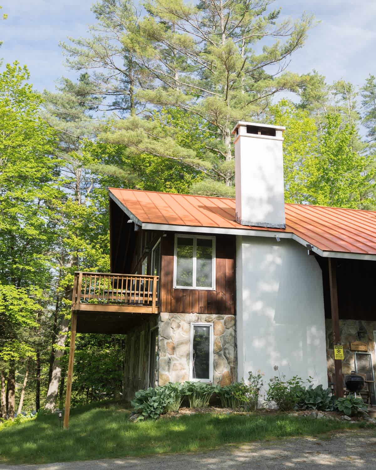 Side view of a rustic chalet-style house with a red metal roof, stone and wood siding, a white chimney, and a wooden balcony, surrounded by lush green trees.