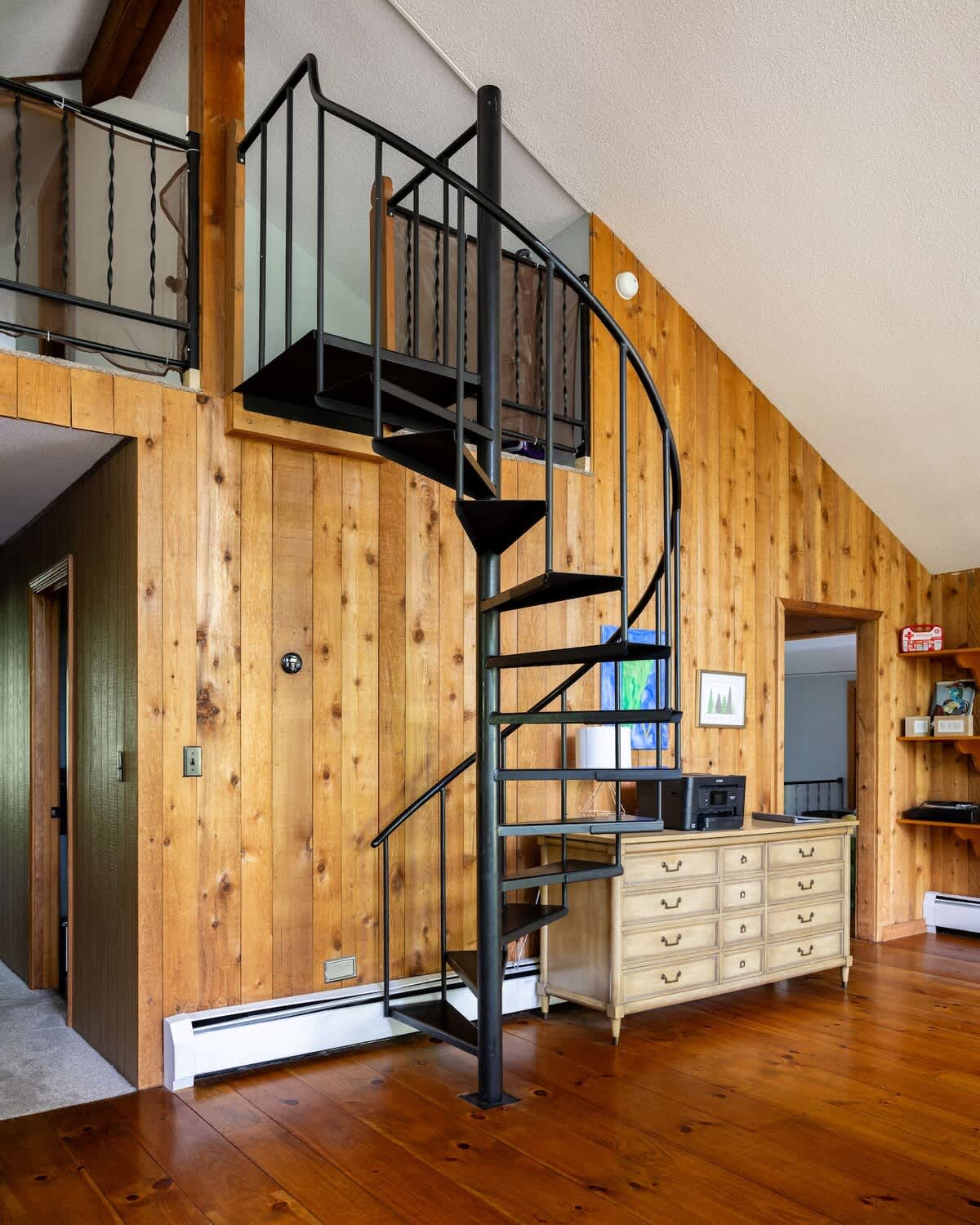 Detailed view of a black metal spiral staircase ascending against a warm wood-paneled wall, leading to an upper-level balcony. A light-colored dresser with multiple drawers is visible below on hardwood floors.