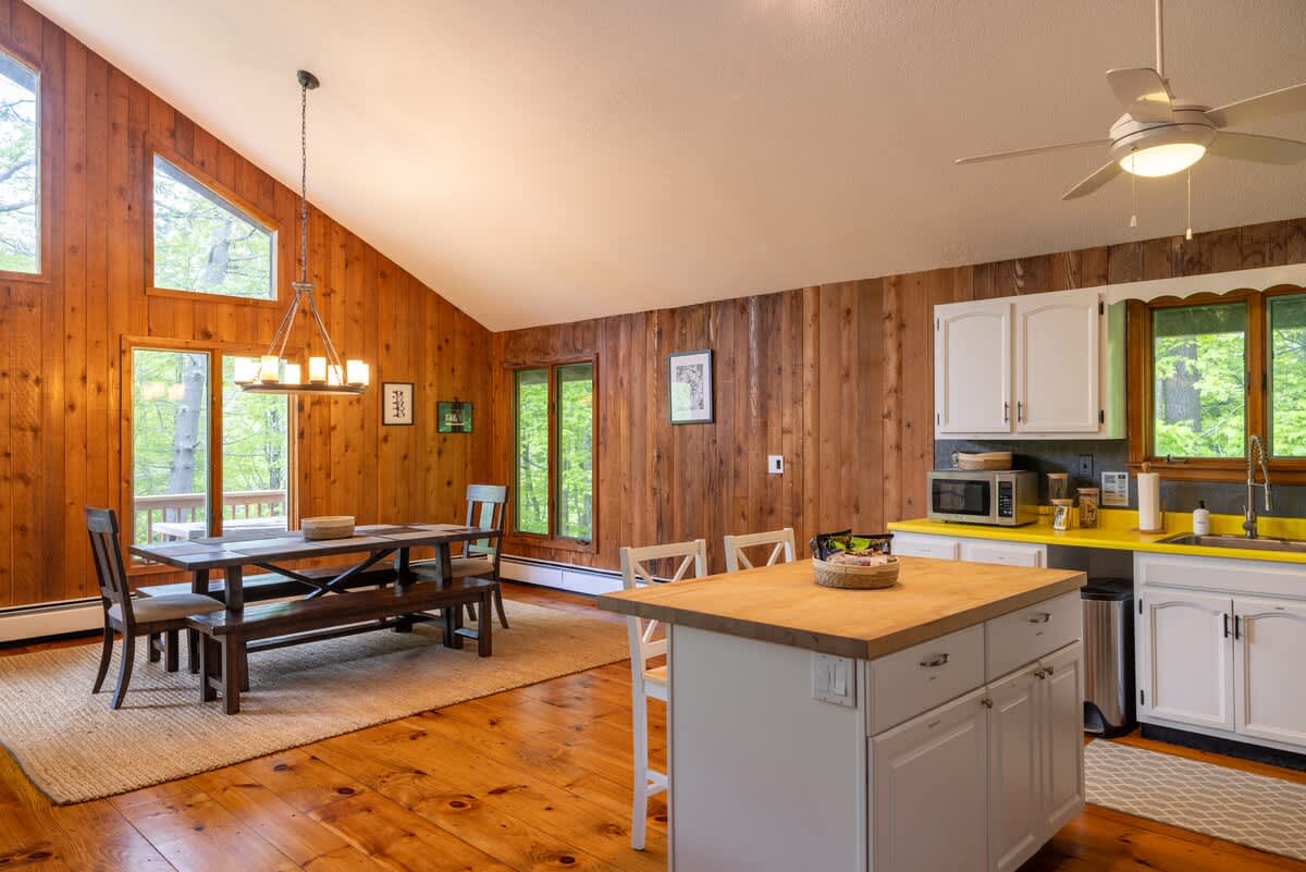 Bright and airy dining space with a vaulted ceiling and large windows offering views of the surrounding trees. A rustic wooden dining table and kitchen island are central to the open layout with wood-paneled walls.