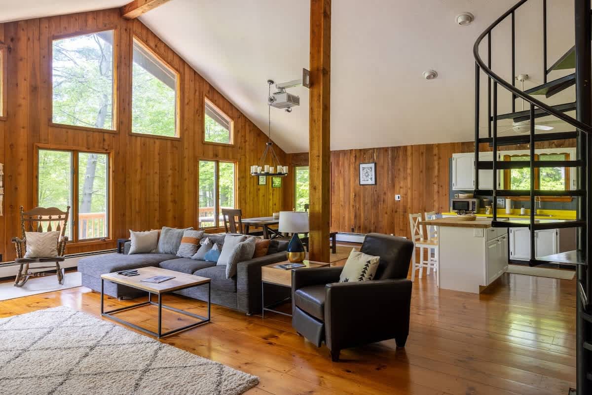 Expansive open-concept living area with vaulted ceilings, large triangular windows, and warm wood paneling, featuring a grey sectional sofa, a dining table, and a black spiral staircase leading to an upper level.
