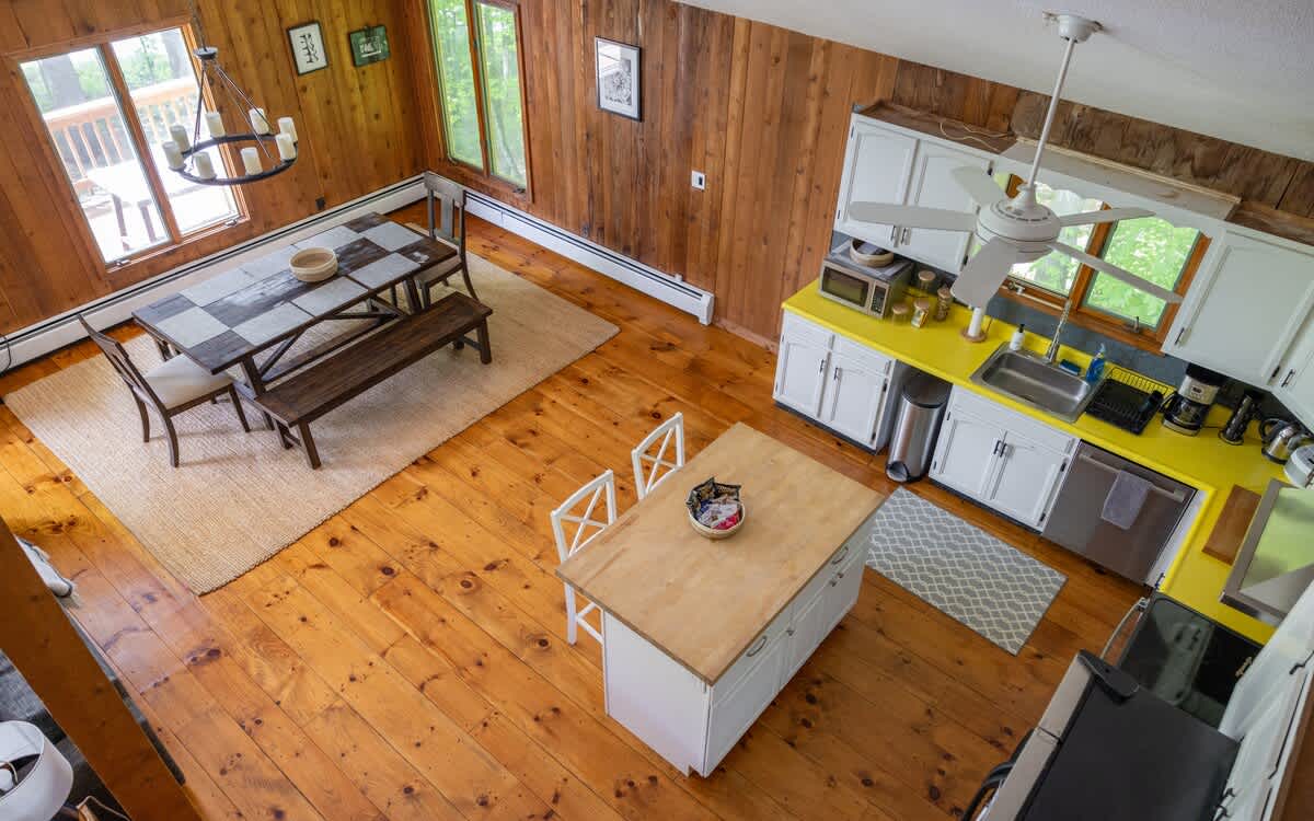 Overhead view of the open-plan kitchen and dining area, highlighting the warm hardwood floors, wood-paneled walls, and the connection between the kitchen island and the dining table. Large windows offer natural light.