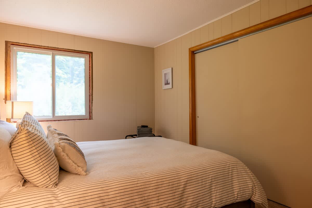Cozy bedroom with light beige walls, a queen-sized bed with striped bedding, a large window, and a sliding closet door, featuring simple wooden trim.
