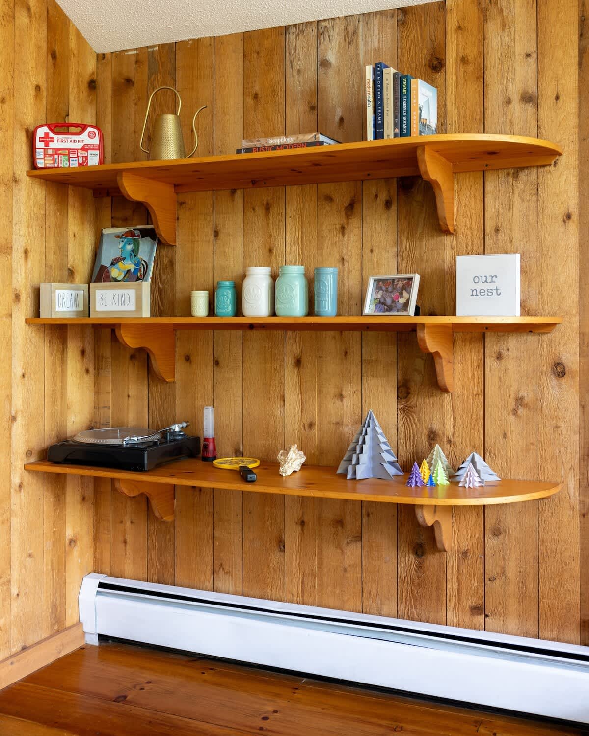Three wooden shelves mounted on a rustic wood-paneled wall, displaying decorative items like jars, a record player, and small geometric sculptures. A white baseboard heater is visible below.