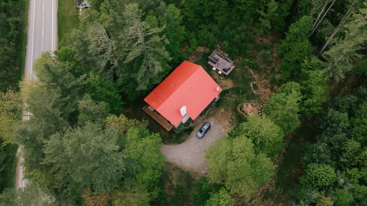 Overhead drone shot of a red-roofed house nestled in a dense green forest, with a car parked in the gravel driveway and a narrow road visible on the left.
